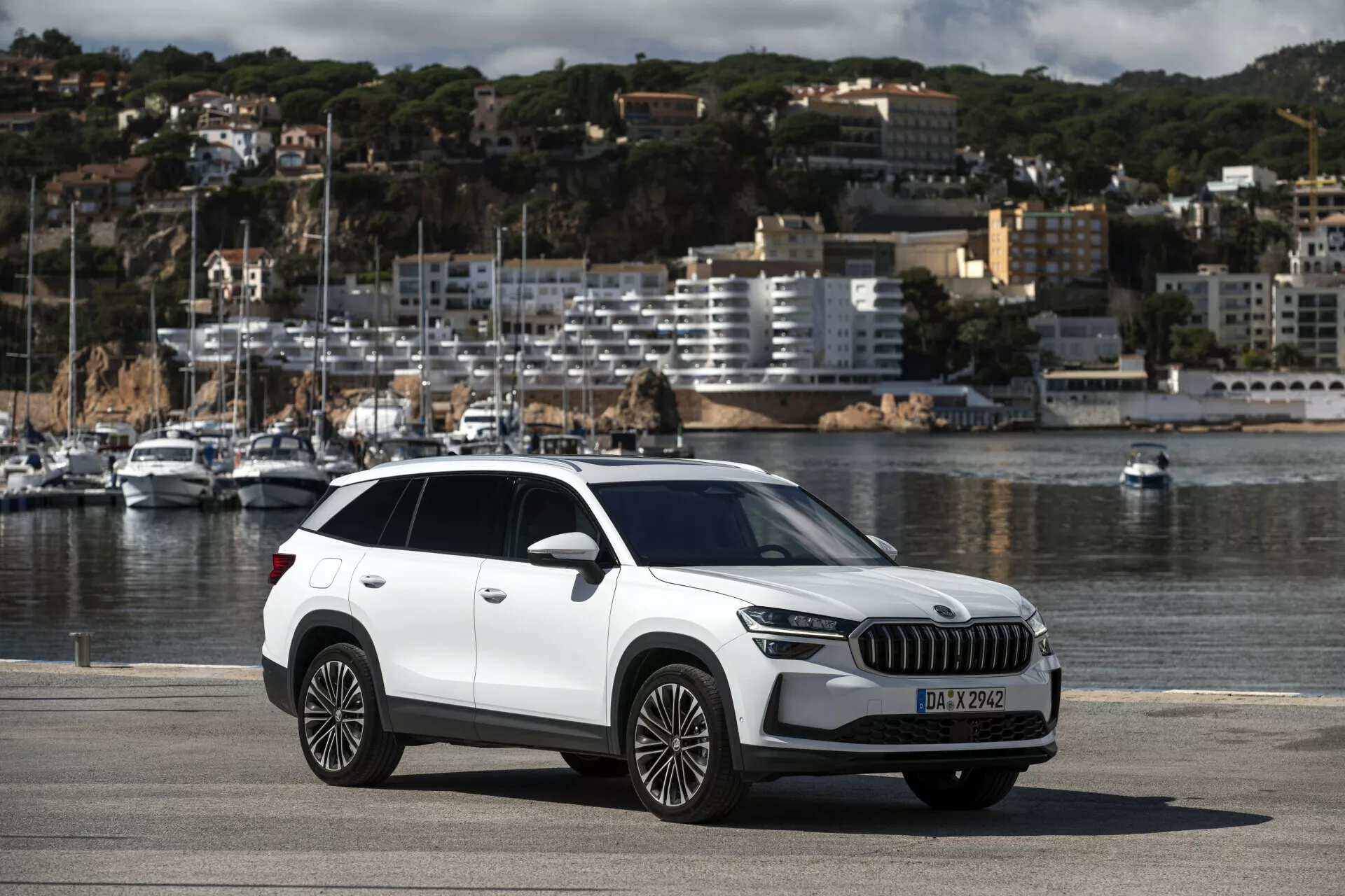 Crossover SUV Skoda Kodiaq 2024 zaparkowany na nadmorskim promenadzie prezentujący pełny widok na samochód w tym nowoczesne elementy stylu jak reflektory LED, duża atrapa chłodnicy, felgi aluminiowe wieloramienne, na tle malowniczego krajobrazu z łodziami i miastem