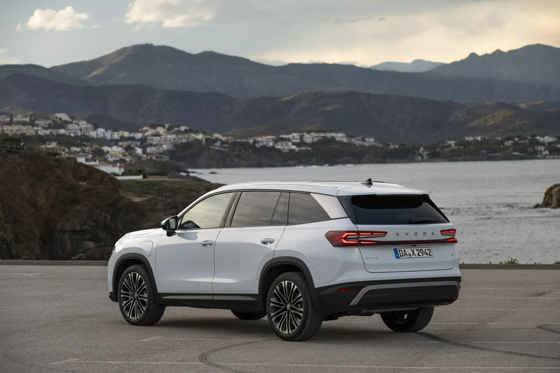 Widok z tyłu trzy czwarte Skoda Kodiaq 2024 w kolorze białym, prezentujący boczne i tylne linie wzornicze, wieloramienne felgi aluminiowe, tylne światła LED oraz oznaczenie modelu, zaparkowany na zewnątrz na drodze przybrzeżnej o złotej godzinie