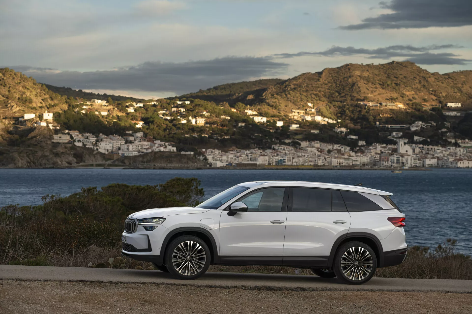 Nowoczesny SUV Skoda Kodiaq 2024 zaparkowany przy nadmorskiej drodze z malowniczym widokiem na zatokę i domy na wzgórzu w tle, prezentujący elegancki design, długość rozstawu osi, dużą prześwit, ostre linie nadwozia, zapadane klamki drzwi, przyciemniane tylne szyby, relingi dachowe, duże koła ze stopu, światła LED, lusterka boczne z kierunkowskazami i panoramiczny dach, wszystko na kontrastującym białym wykończeniu zewnętrznym.