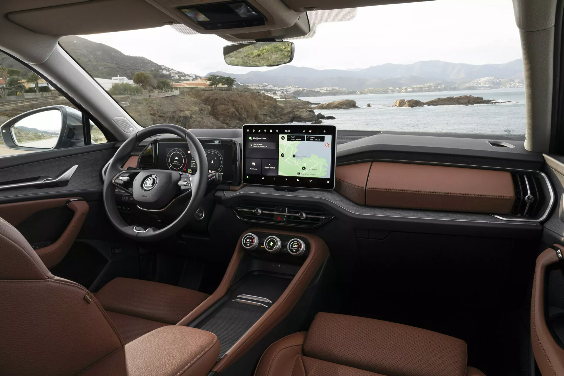 Wnętrze Skoda Kodiaq 2024 z perspektywy pasażera ukazujące deska rozdzielcza, wielofunkcyjna kierownica z logiem, cyfrowy zegar, duży system infotainment z ekranem dotykowym i GPS, luksusowe brązowe skórzane siedzenia i elegancki design konsoli środkowej, z widokiem na spokojny krajobraz nadmorski za oknami