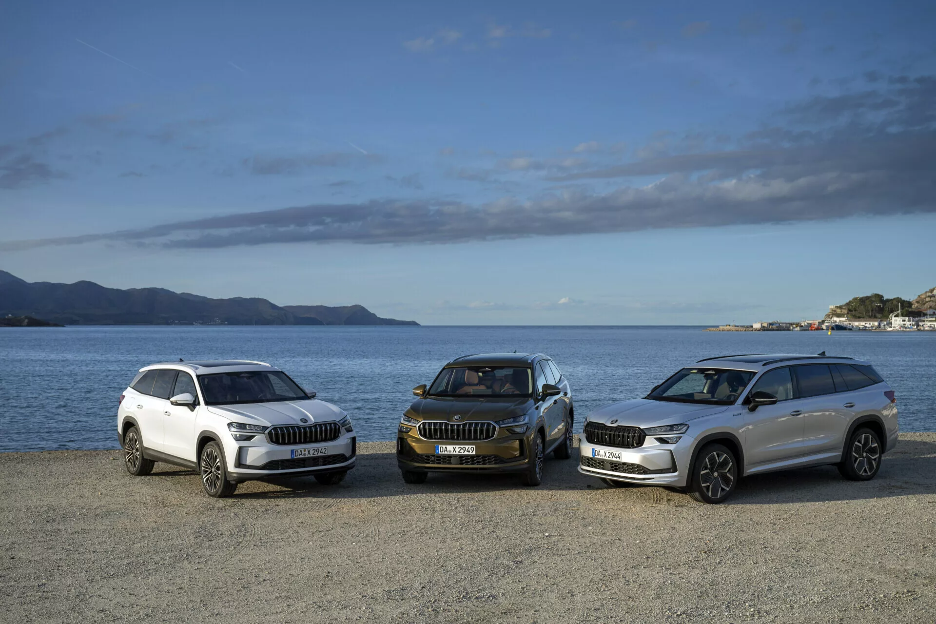 Linia SUV-ów Skoda Kodiaq 2024 w kolorach białym i ciemnym, widok z przodu z boku, zaparkowana przy wodzie z górami i częściowo pochmurnym niebem w tle