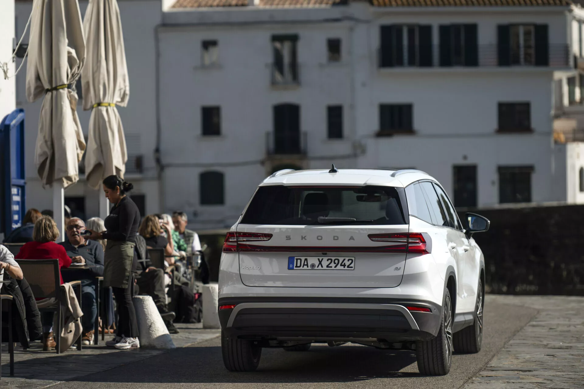Biały Škoda Kodiaq 2024 sfotografowany z tylu, z lekko podwyższonej perspektywy, prezentujący cały tylny koniec, tylne światła, klapę bagażnika, tylną szybę z wycieraczką i dach z anteną i relingami, zaparkowany na brukowanej ulicy przed kawiarnią z klasyczną europejską architekturą w tle.