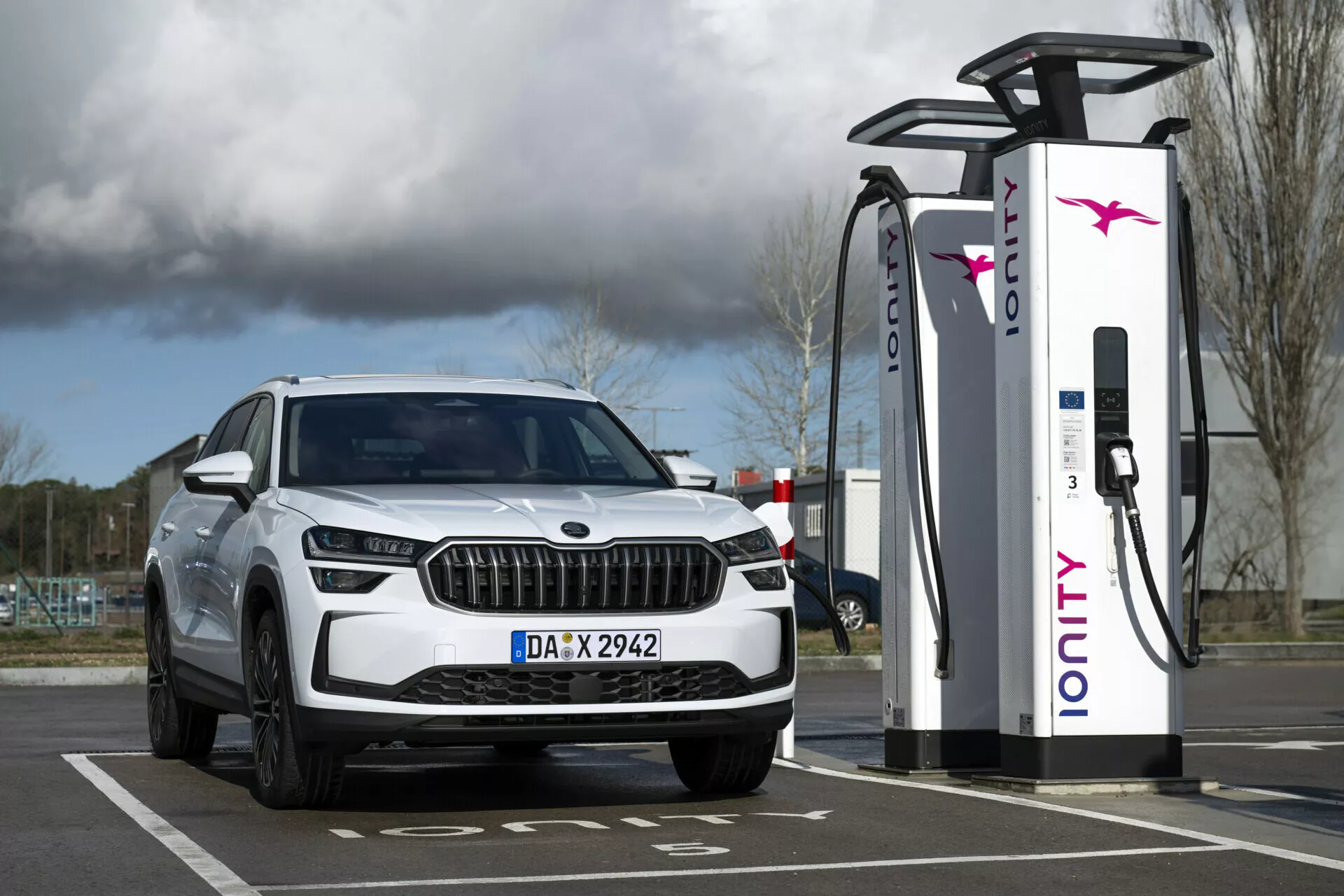 Biały SUV Skoda Kodiaq 2024 zaparkowany przy stacji ładowania elektrycznego, przedstawiający widok z przodu na pierwszy kwartał z reflektorami LED i światłami do jazdy dziennej, podkreślający jego elektryczne lub hybrydowe możliwości oraz luksusowy design grilla.