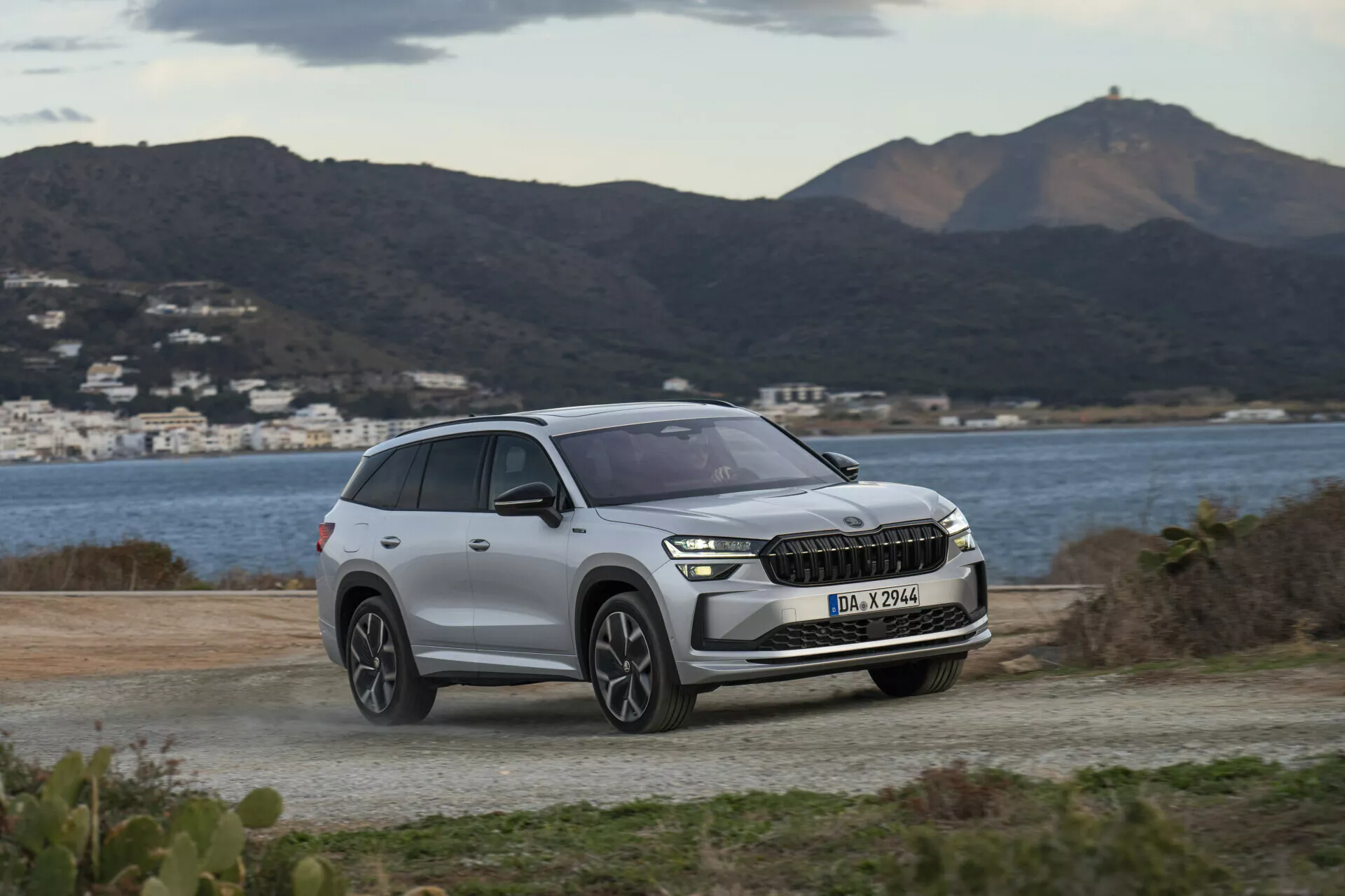 SUV Skoda Kodiaq 2024 zaparkowany na nieutwardzonej powierzchni z widokiem na malownicze wzgórza i akwen, podkreślający nowoczesne reflektory LED pojazdu, przedni grill z potencjalną integracją ADAS i współczesny design kół pod delikatnym, pochmurnym światłem dziennym.