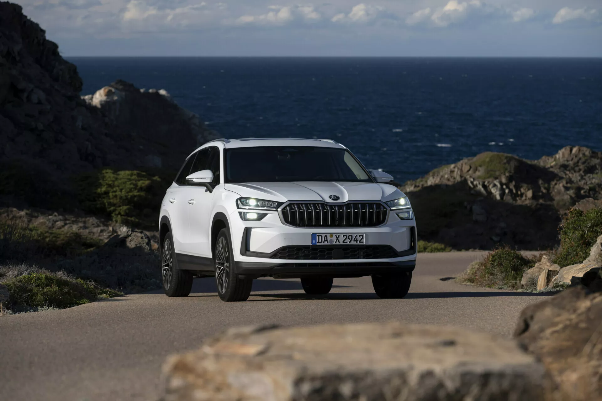Biały SUV Skoda Kodiaq 2024 zaparkowany na nadmorskiej drodze z oceanem w tle, cechujący się wyróżniającą się przednią kratą z pionowymi listwami przypominającymi chrom, diodami LED do jazdy dziennej, kołami ze stopu o wielu szprychach i szczegółowym widokiem z przodu z trzech czwartych