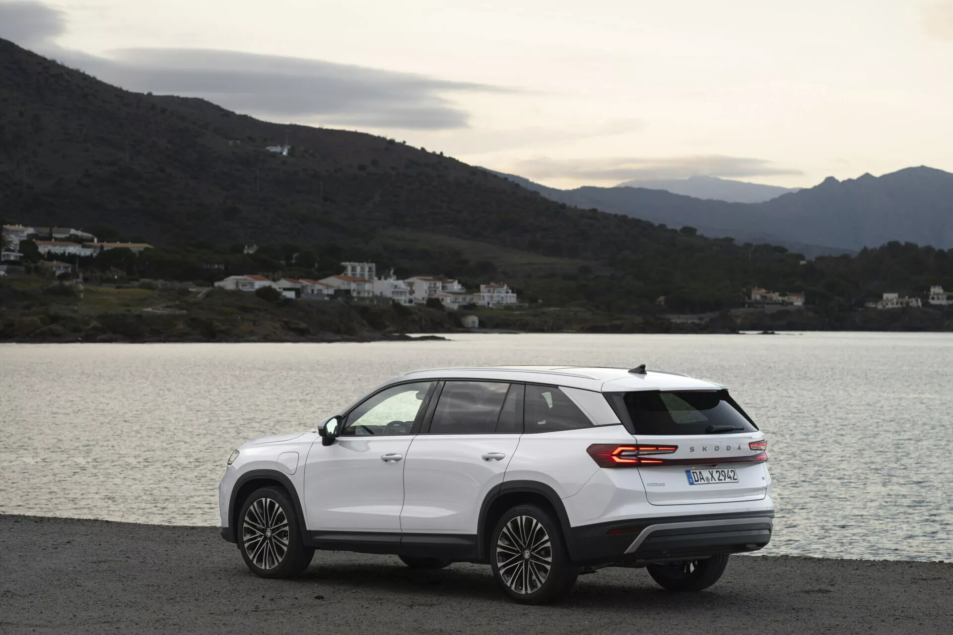 Nowoczesny SUV Skoda Kodiaq 2024 zaparkowany bokiem na malowniczej przybrzeżnej drodze, z aerodynamicznym designem, pasem świateł z tyłu i felgami aluminiowymi o wielospiczastym wzorze, w pochmurny dzień z rozproszonym naturalnym oświetleniem, z tłem wody, wzgórz i budynków