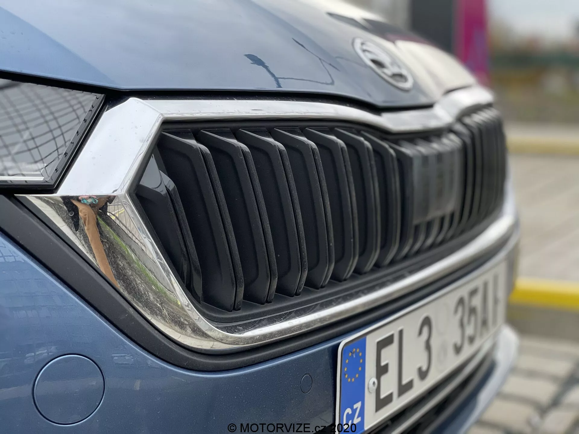 Zbliżenie przedniej części samochodu, skupiające się na grillu i reflektorze. Zdjcie wykonane pod przednim prawym kątem, lekko w górę, pokazuje szczegółowo prawą stronę grilla, prawy reflektor i część maski. Pionowe listwy grilla są wyraźne, co wskazuje na odważny design. Skomplikowany reflektor sugeruje zaawansowaną technologię oświetleniową, taką jak LED lub systemy adaptacyjne. Maska jest lekko rozmyta, podkreślając teksturę grilla i reflektora. Samochód jest dobrze utrzymany, z odblaskami wskazującymi na ustawienia zewnętrzne w pochmurny dzień lub w cieniu.