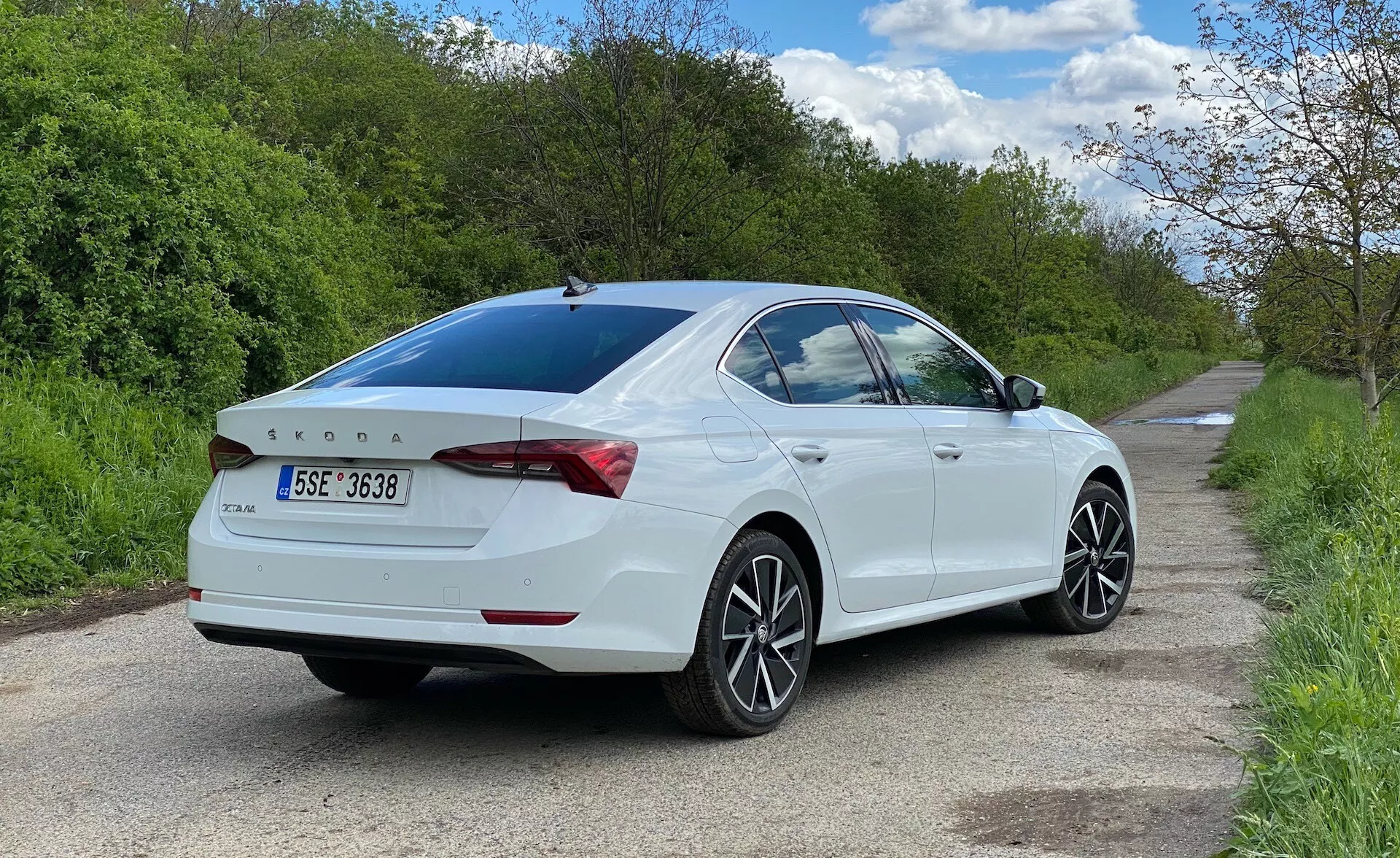 Widok tylny na trzy czwarte białego sedana Skoda Octavia 2020 zaparkowanego po prawej stronie wąskiej drogi, ukazujący bok pasażera z poziomu gruntu. Samochód wypełnia większość kadru, podkreślając linie projektowe, tylne światła otaczające bagażnik, subtelny zintegrowany spojler, odznakę tylną, wieloszprychowe felgi aluminiowe, lekko przyciemniane tylne okno i lusterka boczne z wbudowanymi kierunkowskazami.