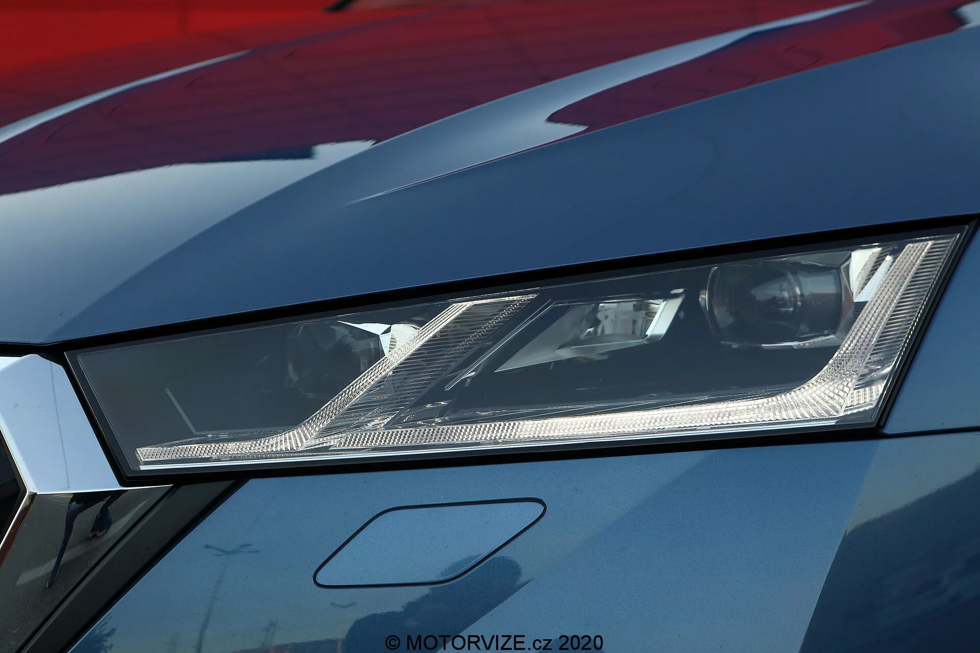 Zbliżenie lewego reflektora w modelu Škoda Octavia 2020 limuzyna, podkreślające jego kanciasty design i nowoczesną technologię oświetlenia LED lub laserowego. Fotografia, wykonana z niskiego kąta, pokazuje skomplikowane wewnętrzne reflektory i drobne szczegóły wewnątrz reflektora pokrytego przezroczystą soczewką. Obraz o wysokiej rozdzielczości ujawnia również część maski i błotnika z metalicznym wykończeniem lakieru, sugerując pojazd wyższej klasy lub dobrze utrzymany. Ten obraz podkreśla zaawansowane oświetlenie samochodowe i agresywny styl kojarzony ze sportowymi samochodami.