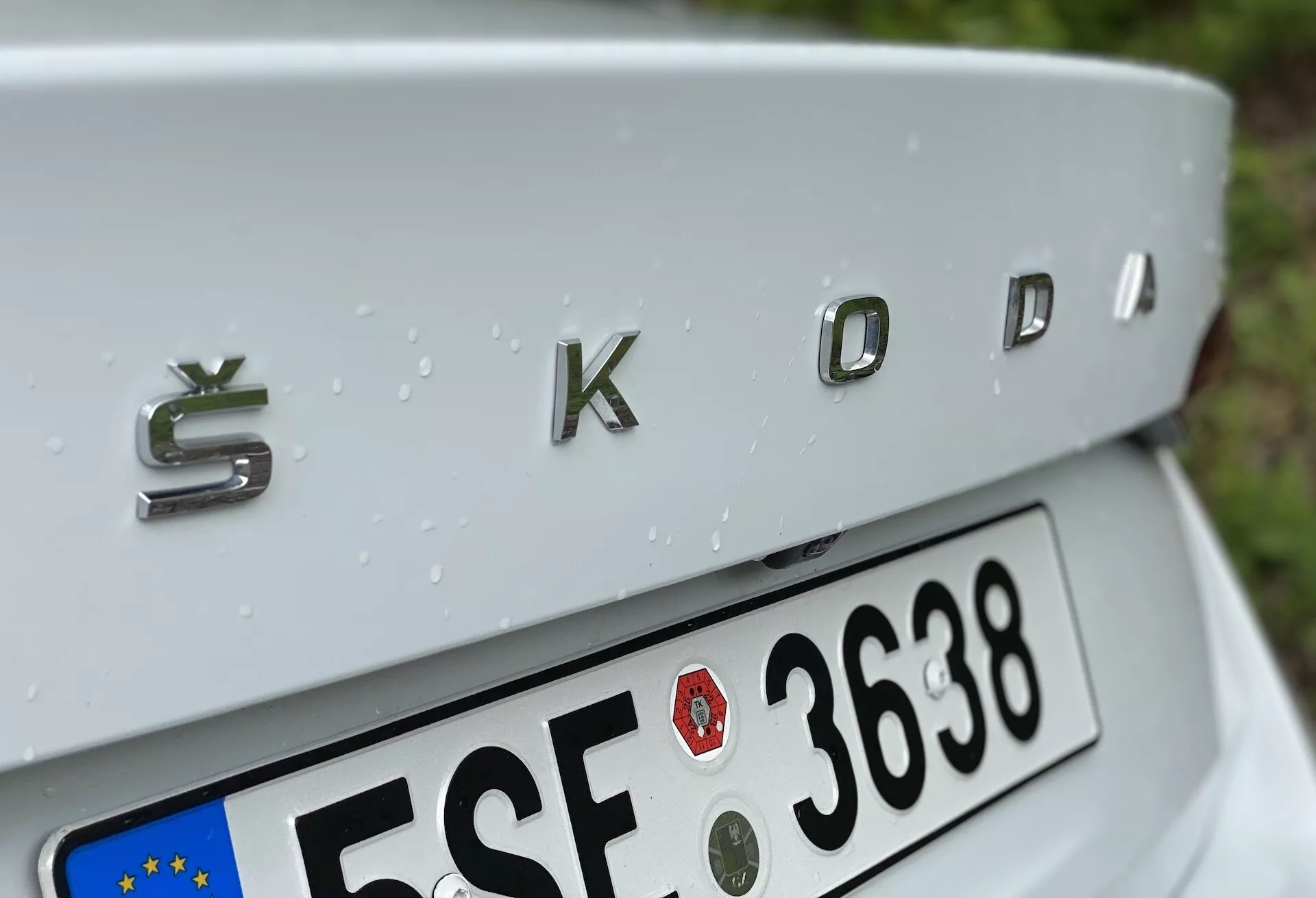 Zbliżenie tylnej części Škoda Octavia 2020 (octavia-limousine) z naciskiem na chromowane emblematy 'ŠKODA' z trójwymiarowym metalicznym wykończeniem. Ujęcie lekko z góry pokazuje delikatnie opadającą pokrywę pomaznika, częściowo widoczną tablicę rejestracyjną w stylu UE poniżej oraz krople deszczu zdobiące srebrną powierzchnię samochodu, wskazujące na niedawno spadły deszcz.