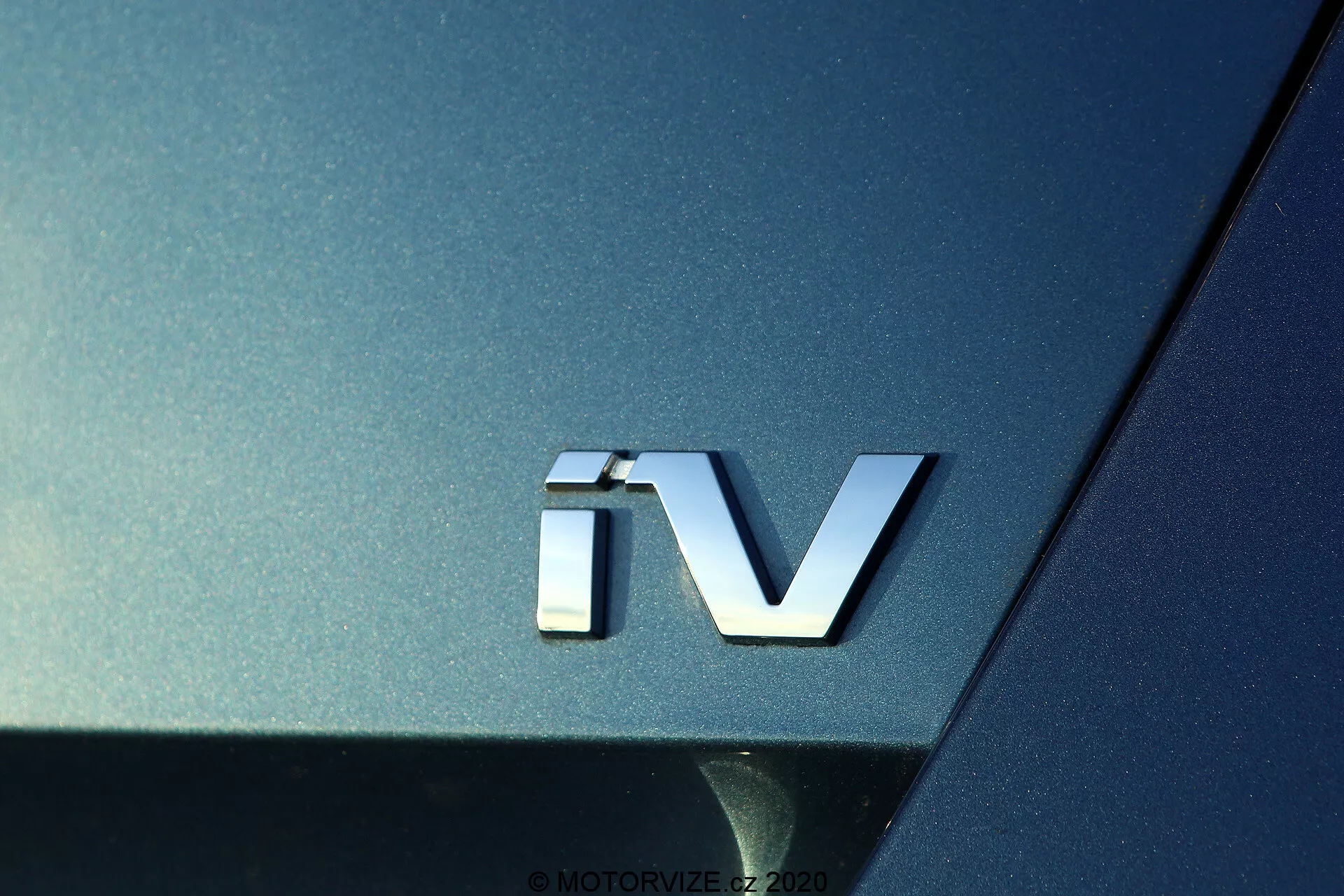 Zbliżenie emblematu 'iV' Skoda Octavia 2020, oznaczającego zelektryfikowany układ napędowy, ze srebrnym trójwymiarowym znaczkiem na ciemnym metalicznym nadwoziu, podkreślającym zakrzywioną powierzchnię i odblaskowe właściwości tylnej części pojazdu.