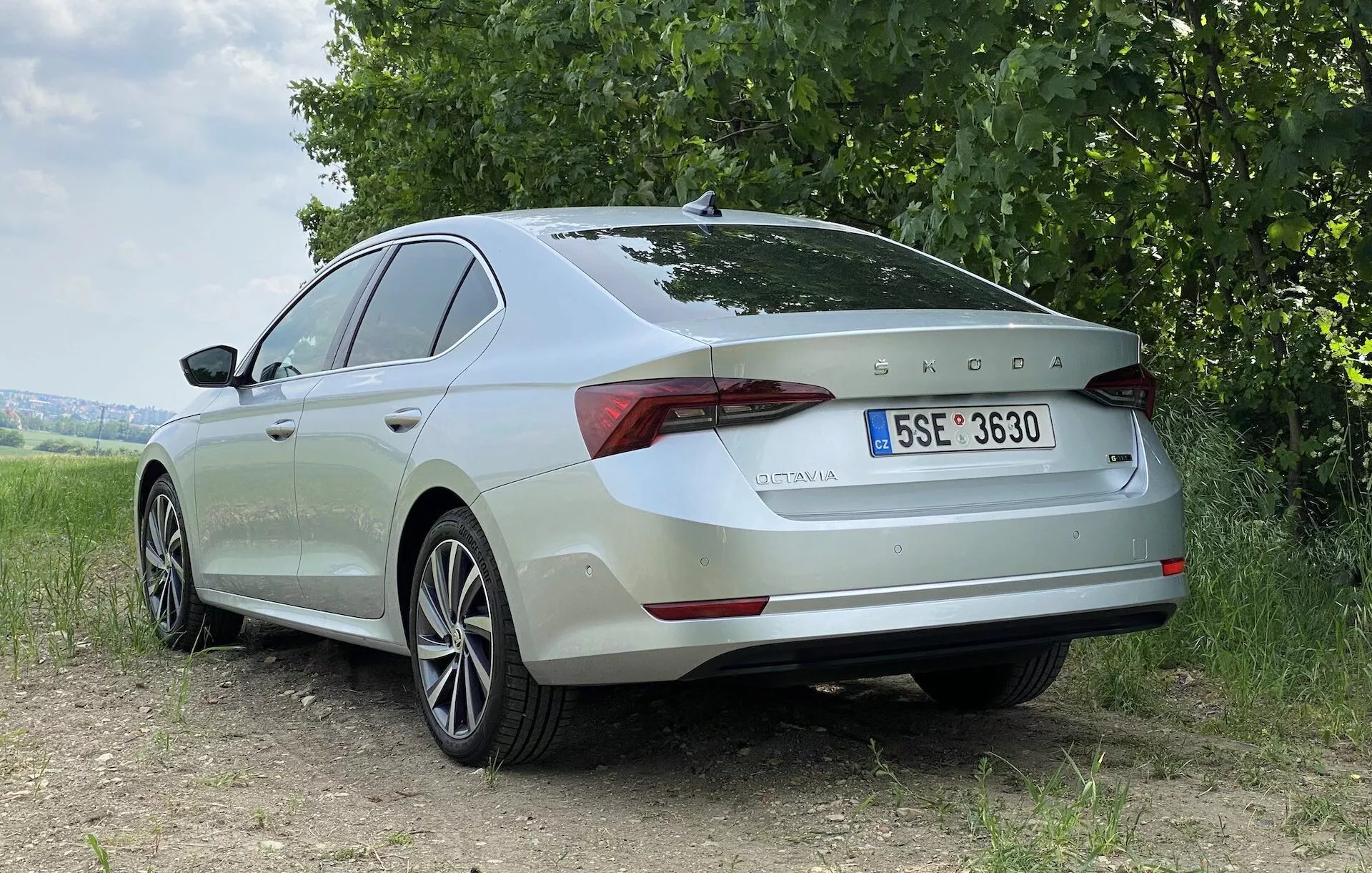 Tylny widok z trzech czwartych jasnego sedana Skoda Octavia 2020 na ziemistej powierzchni, z trawą i drzewami w tle. Zdjęcie podkreśla tylny i boczny profil pojazdu, pokazując gładkie linie, konturowane powierzchnie, aluminiowe felgi i eleganckie tylne światła LED. Emblemat Skoda i nazwa modelu 'OCTAVIA' są wyraźnie widoczne na pokrywie bagażnika, wraz z europejską tablicą rejestracyjną. Naturalne, miękkie światło podkreśla elementy stylistyczne samochodu bez ostrych cieni, co sugeruje zachmurzone niebo lub rozproszony promień słoneczny.