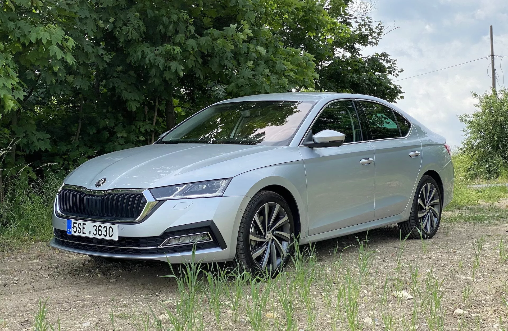 Skoda Octavia 2020 widziana z niskiego przedniego kąta trzy czwarte, prezentująca szczegółowy lewy przedni bok, w tym zestaw reflektorów LED, wyraźną kratkę, przedni zderzak z czujnikami, rzeźbione linie maski, wieloramienne felgi aluminiowe, zintegrowany kierunkowskaz w lusterku bocznym i lekko przyciemniane szyby, umieszczona na nieutwardzonej powierzchni z zielonymi liśćmi i drucianym ogrodzeniem w wiejskim lub półwiejskim otoczeniu.