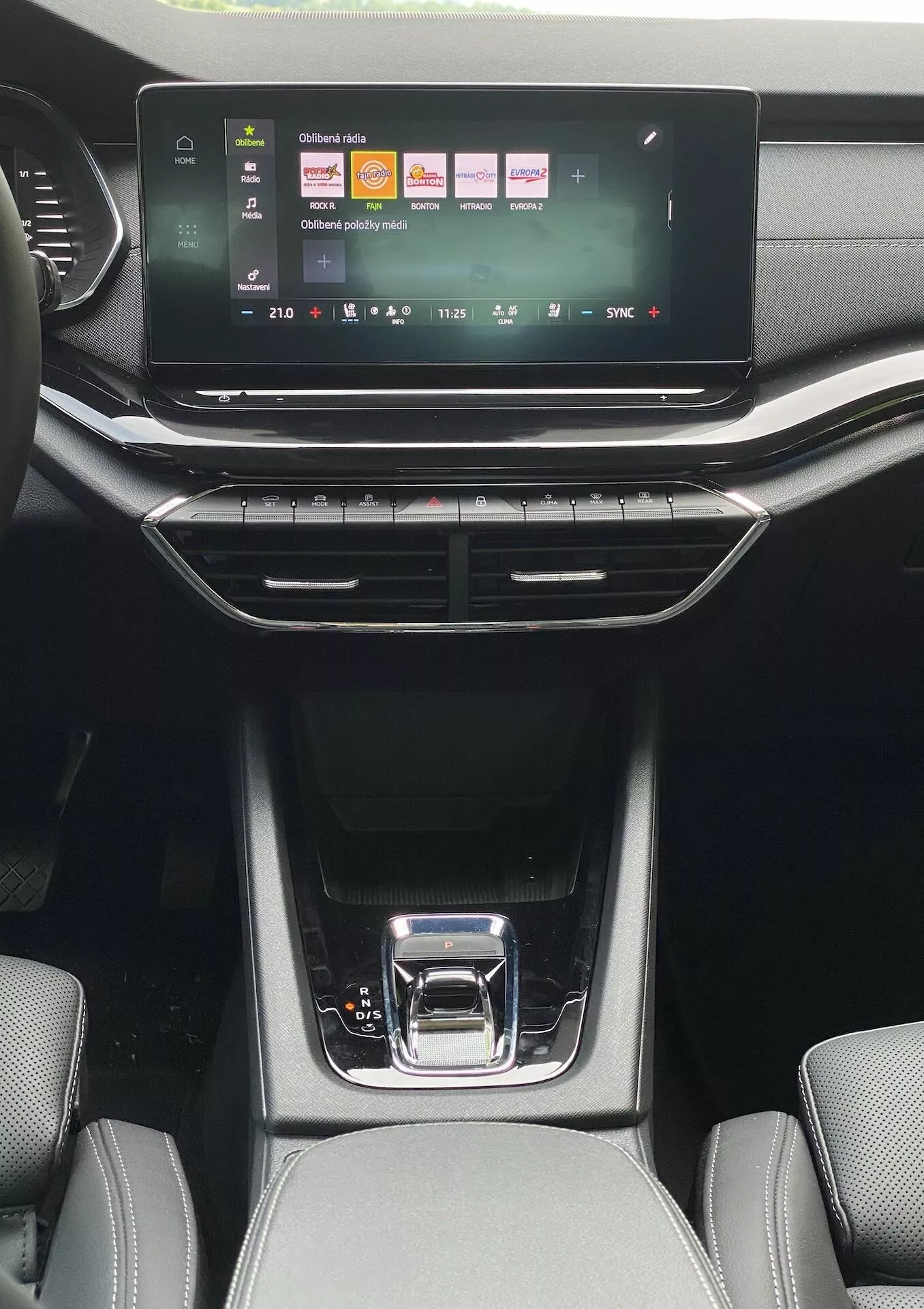 Widok wnętrza z tyłu pasażerskiego Skody Octavia limousine z 2020 roku, prezentujący system informacyjno-rozrywkowy z kolorowymi ikonami dla mediów, połączeń telefonicznych i ustawień pojazdu. Poniżej znajdują się przyciski klimatyzacji dwustrefowej z cyfrowym wyświetlaczem temperatury, otoczonym pionowymi i poziomymi wylotami powietrza. Elegancka konsola środkowa ma błyszczącą czarną dźwignię zmiany biegów ze srebrnym uchwytem, schowki z gumowymi matami oraz widoczną teksturowaną tapicerkę przednich siedzeń w naturalnym świetle dziennym.