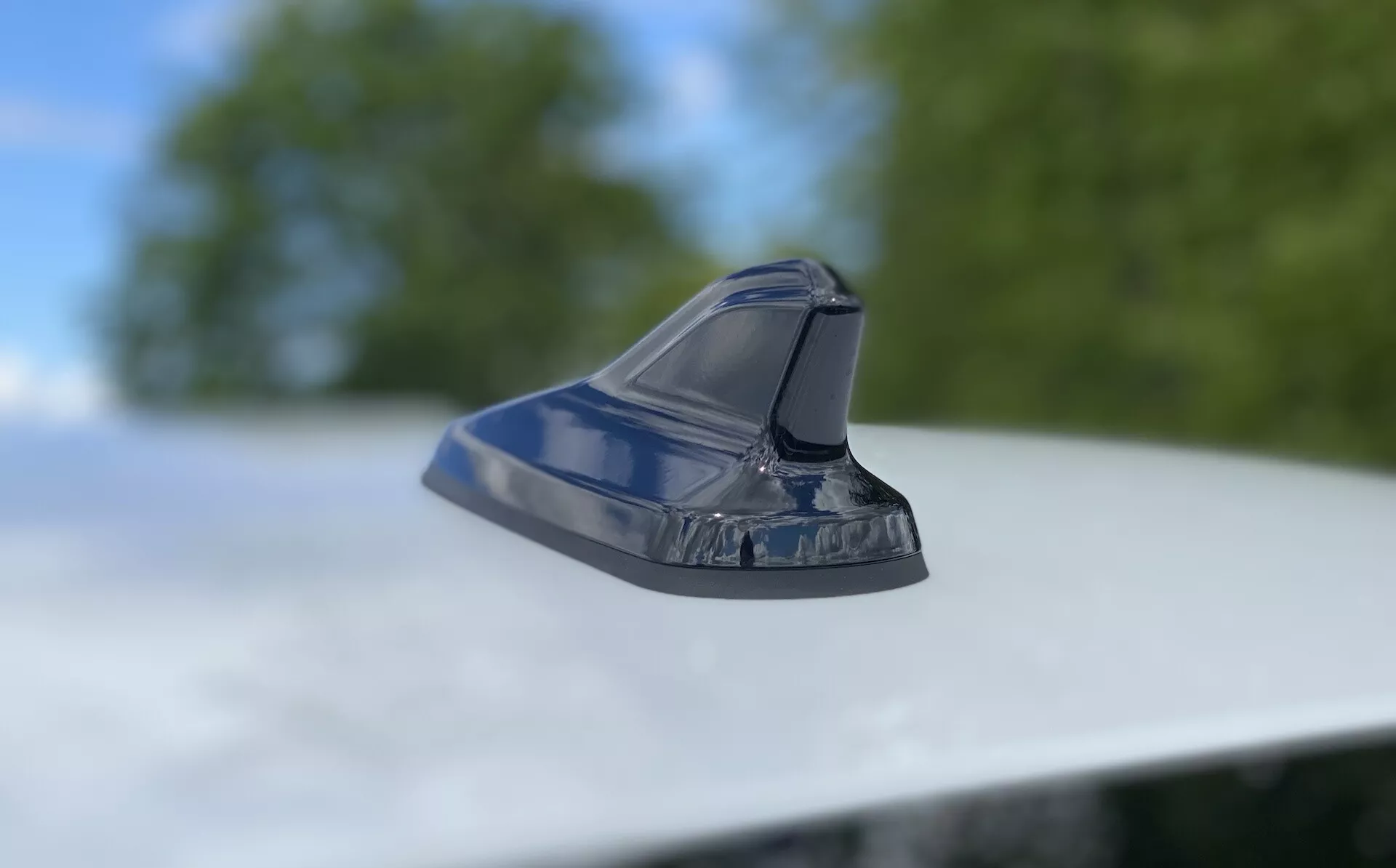 Zbliżenie anteny w stylu płetwy rekina na dachu pojazdu, prezentujące nowoczesny design motoryzacyjny. Zdjęcie wykonane z niskiego kąta z aparatem lekko pochylonym w górę, skupiające się na lśniącej, aerodynamicznej antenie na rozmazanym tle liści i nieba. Opływowy kształt sugeruje funkcje, takie jak radio FM/AM, nawigacja satelitarna i zaawansowane technologie komunikacyjne. Metaliczne wykończenie odzwierciedla dobrze utrzymany wygląd zewnętrzny pojazdu.