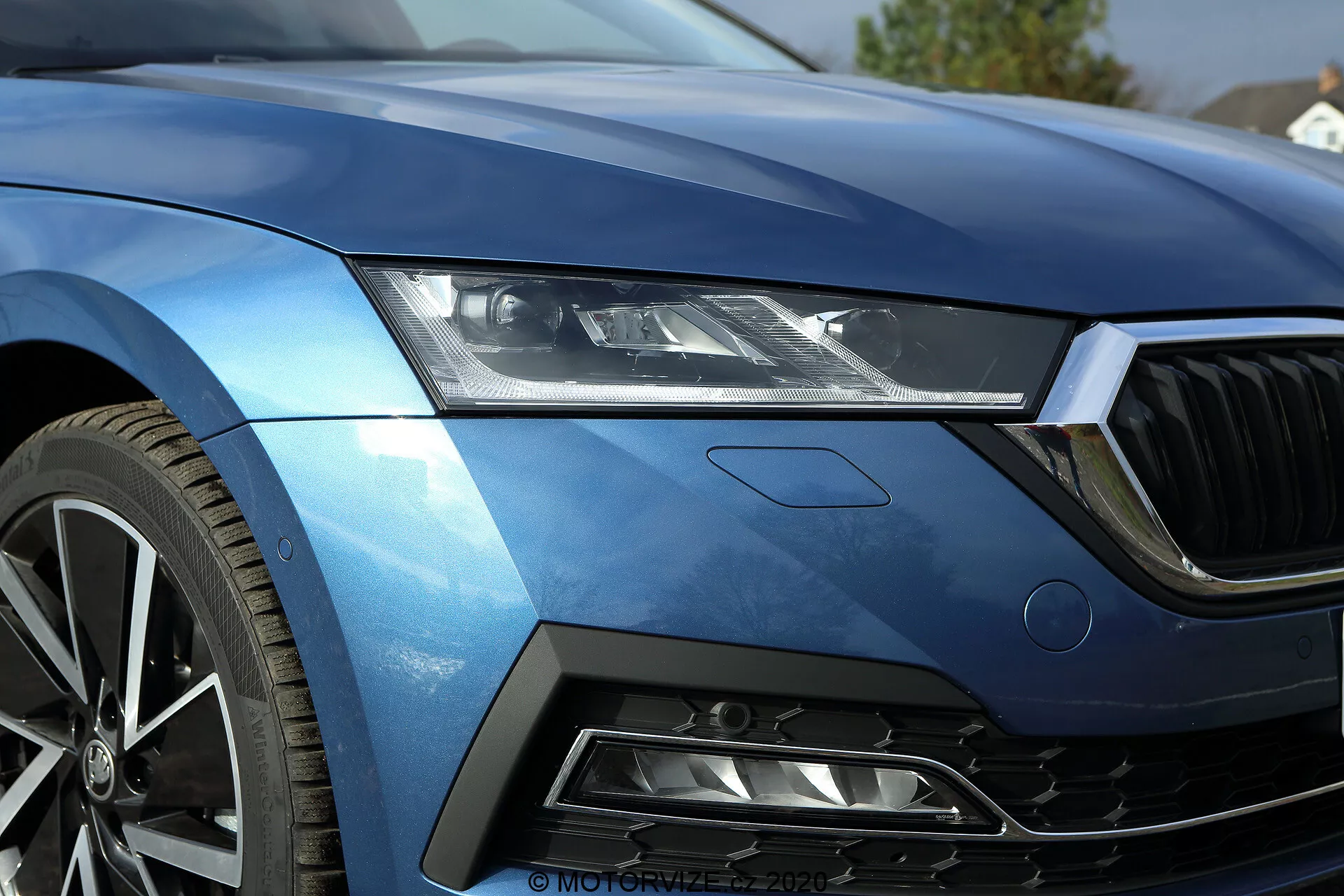 Zbliżenie przedniej lewej ćwiartki limuzyny Skoda Octavia 2020, ukazujące elegancki zestaw reflektorów LED, część maski, lewą krawędź przedniego grilla, aerodynamiczny przedni lewy błotnik oraz segment zderzaka z wlotem powietrza w kształcie sześciokątnej siatki. Metaliczny niebieski lakier i ostre linie podkreślają luksus i sportowy charakter. Wieloramienna felga aluminiowa sugeruje wysoką wydajność. Wzornictwo zawiera subtelny znaczek, wskazujący na określony poziom wyposażenia lub pakiet technologiczny. Ten obraz pięknie uchwyci zaawansowaną technologię oświetlenia pojazdu, wykończenie lakieru oraz cechy wzornictwa skoncentrowanego na wydajności.
