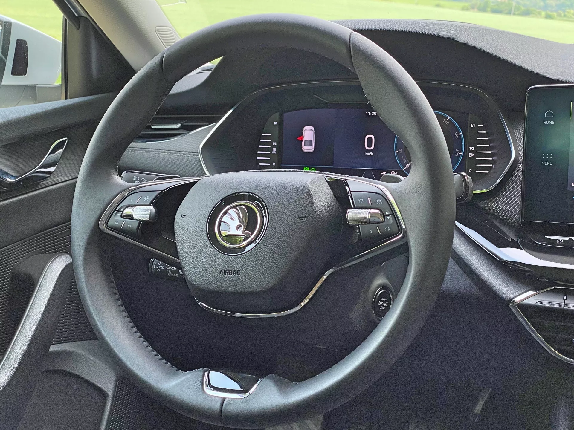 Wnętrze Skody Octavia Limousine 2020 z perspektywy kierowcy, podkreślające nowoczesną, trójramienną kierownicę obszytą skórą z różnymi kontrolkami do regulacji głośności, poleceń głosowych i ustawień tempomatu. Za kierownicą znajduje się w pełni cyfrowy zestaw wskaźników wyświetlający prędkościomierz w stanie spoczynku i wskaźnik otwartych drzwi. Konsola centralna zawiera ekran dotykowy do systemu infotainment do multimediów, nawigacji i ustawień pojazdu. Deska rozdzielcza pokazuje nowoczesny design z wysokiej jakości materiałami.