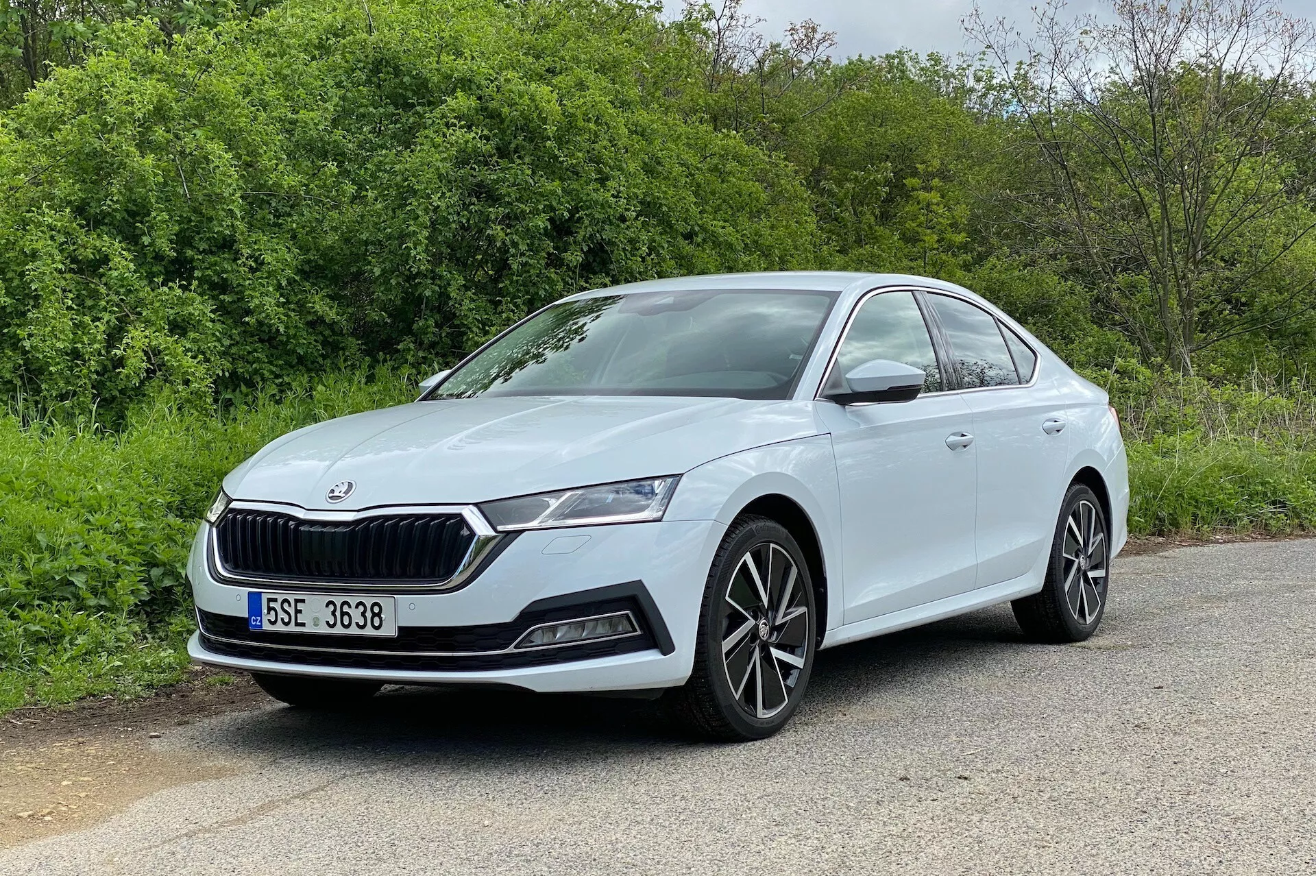 Zdjęcie przedstawia samochód Skoda Octavia 2020 w trójwymiarowym ujęciu z przodu, ukazując przód oraz profil boczny po stronie kierowcy. Samochód, zaparkowany na asfaltowej powierzchni z naturalnymi elementami jak drzewa czy krzewy w tle, charakteryzuje się wyróżniającym się grillem, eleganckimi reflektorami LED, obudowami świateł przeciwmgielnych oraz wlotami powietrza w zderzaku. Wieloramienne aluminiowe felgi oraz gładkie, aerodynamiczne linie boczne podkreślają jego nowoczesny i sportowy wygląd. Dobrze oświetlony i niezasłonięty widok podkreśla kolorowe klamki drzwi oraz widoczną linię charakterystyczną biegnącą przez drzwi, co czyni to doskonałą reprezentacją wzornictwa samochodowego.