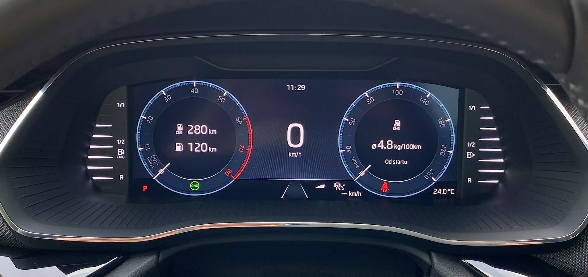Zbliżenie cyfrowego zestawu wskaźników Skoda Octavia 2020 z perspektywy kierowcy, z szerokoekranowym wyświetlaczem o czarnym tle i wyraźną grafiką cyfrową. Lewa strona pokazuje obrotomierz do 8 000 obr./min z czerwonym polem od 6 500 obr./min, wskazówka na biegu jałowym. W centrum wyświetla się prędkość '0 km/h', zegar '11:29', zużycie paliwa '4,8 l/100km' i przebieg '6540 km'. Po prawej stronie znajduje się grafika przypominająca prędkościomierz z oznaczeniami do 280 km/h i wartością temperatury '24°C'. Ikony obejmują kompas nawigacyjny i wskaźnik poziomu paliwa.