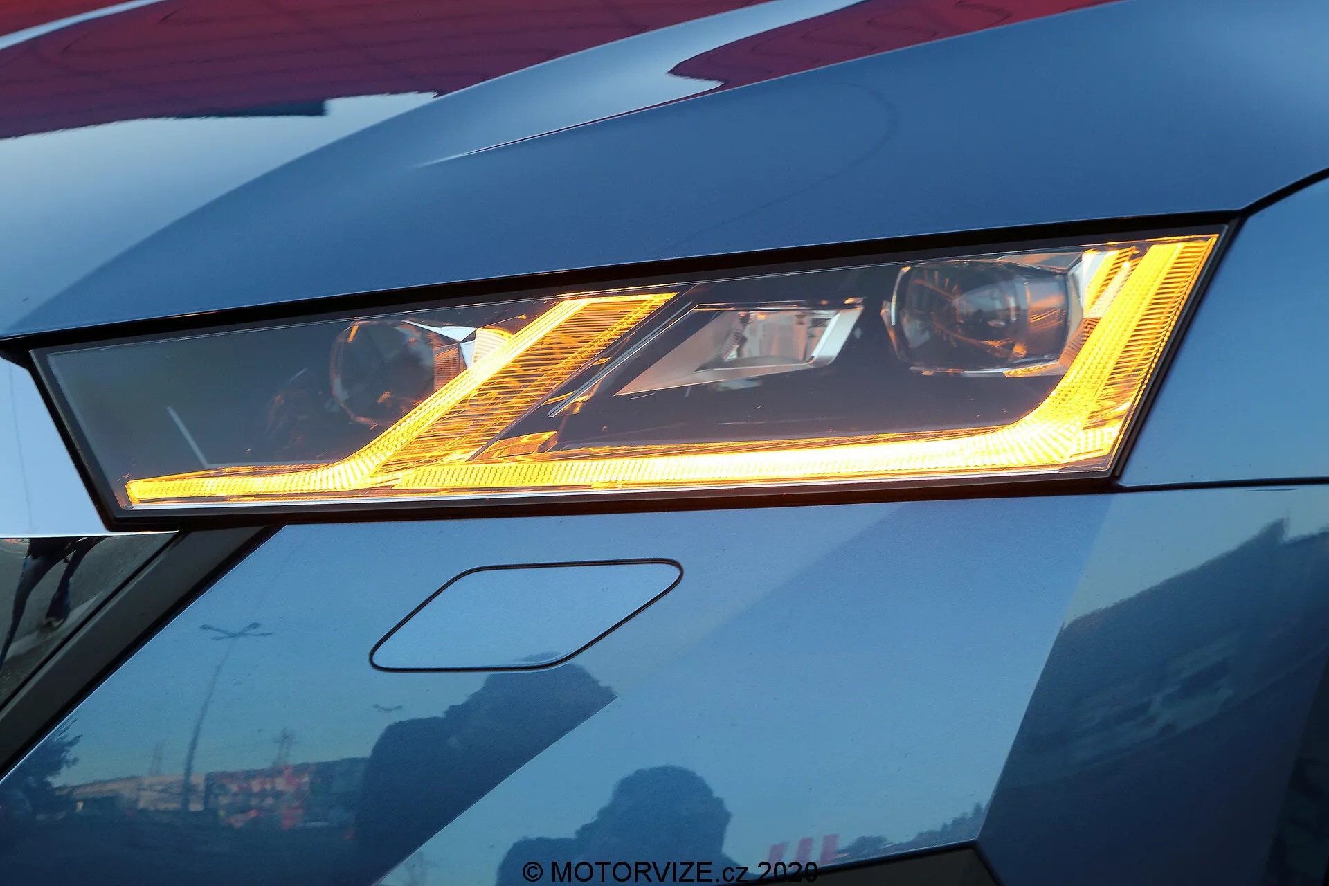 Zbliżenie zaawansowanego reflektora LED ciemnoczerwonej Skody Octavia 2020, podkreślające jego jasne oświetlenie i charakterystyczne wzory świetlne. W zaawansowanym designie pod głównym reflektorem znajduje się dzienne światło (DRL), a elegancka karoseria wyróżnia się ostrymi krawędziami. Odbicia nieba i budynków na dobrze wypolerowanej powierzchni sugerują wykończenie premium. Kadrowanie lekko ujawnia przedni zderzak, skupiając się na innowacjach w projektowaniu motoryzacyjnym.