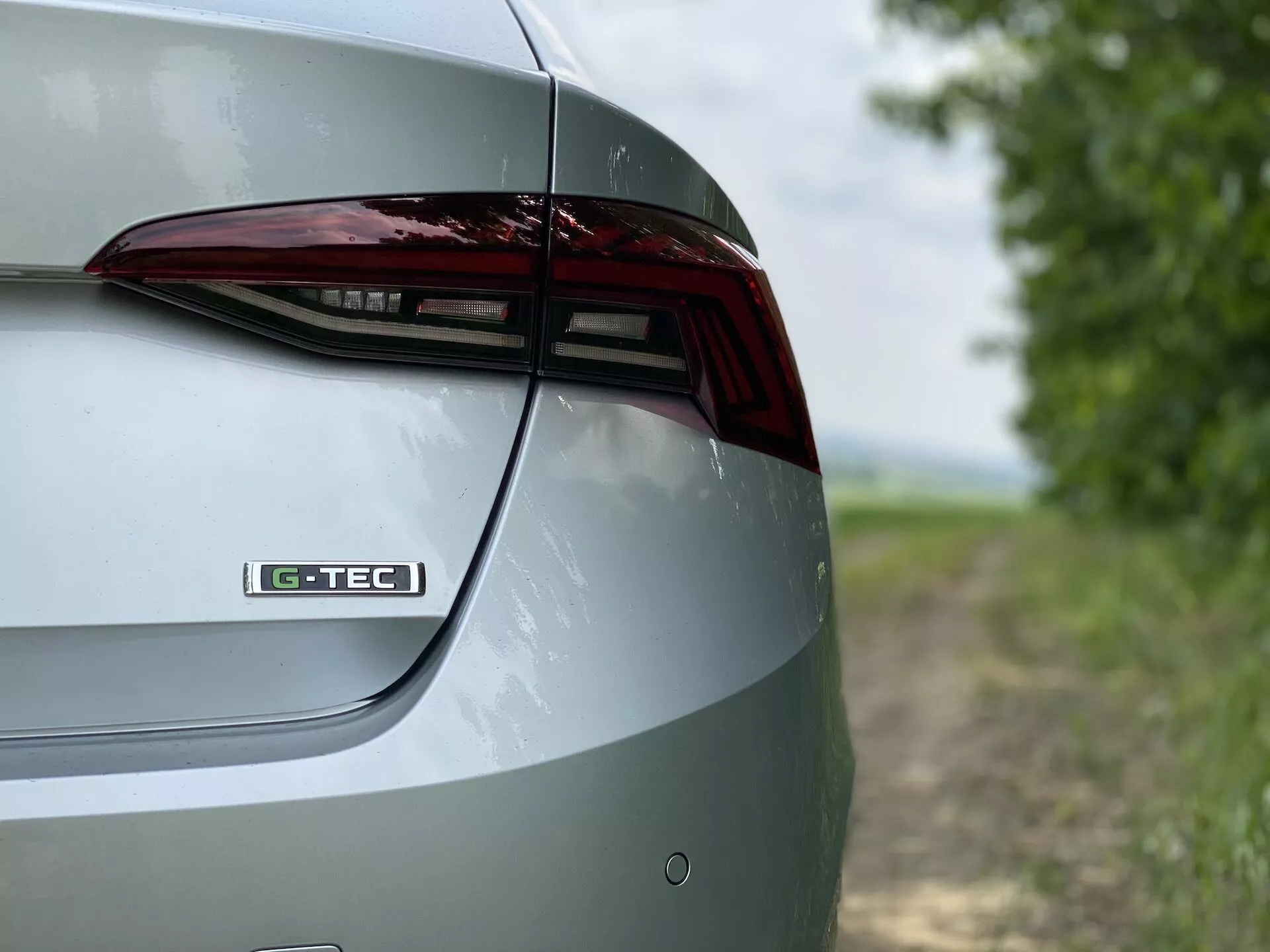 Zbliżony, szczegółowy widok prawej tylnej części samochodu Skoda Octavia 2020 limuzyna. Ujęcie ukazuje elementy projektowe pokrywy bagażnika, tylną lampę LED i część tylnego zderzaka pod skosnym, lekko podwyższonym kątem. Znak 'G-TEC' na pokrywie bagażnika sugeruje specyficzną technologię lub wykończenie. Metaliczne wykończenie lakieru podkreśla wypolerowany wygląd zewnętrzny samochodu. Rozmyte tło tworzy wiejski krajobraz, który podkreśla cechy i design pojazdu.