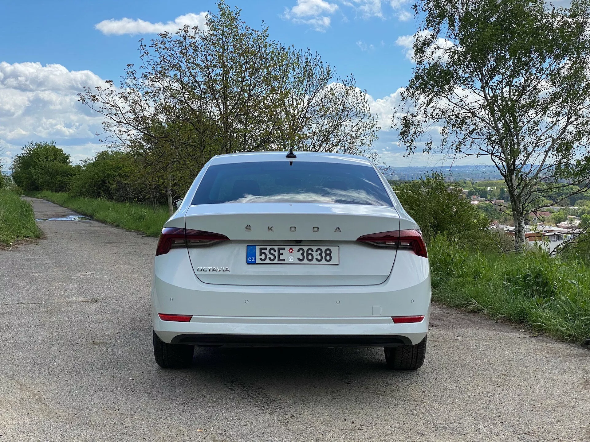 Widok tylnej części białego modelu Škoda Octavia 2020 limuzyna, stojącego na wąskiej asfaltowej drodze w wiejskim otoczeniu. Obrazek, wykonany z poziomu gruntu pod prostym kątem, pokazuje całą tylną część samochodu, w tym pokrywę bagażnika, tylny zderzak, tylne światła, tablicę rejestracyjną, tylną szybę i logo Škody wyraźnie widoczne pośrodku pokrywy bagażnika z napisem 'Octavia' po lewej. W otoczeniu pojazdu znajdują się drzewa i pochmurne niebo, a w tle widoczna jest oddalona miejscowość lub krajobraz poniżej linii horyzontu.