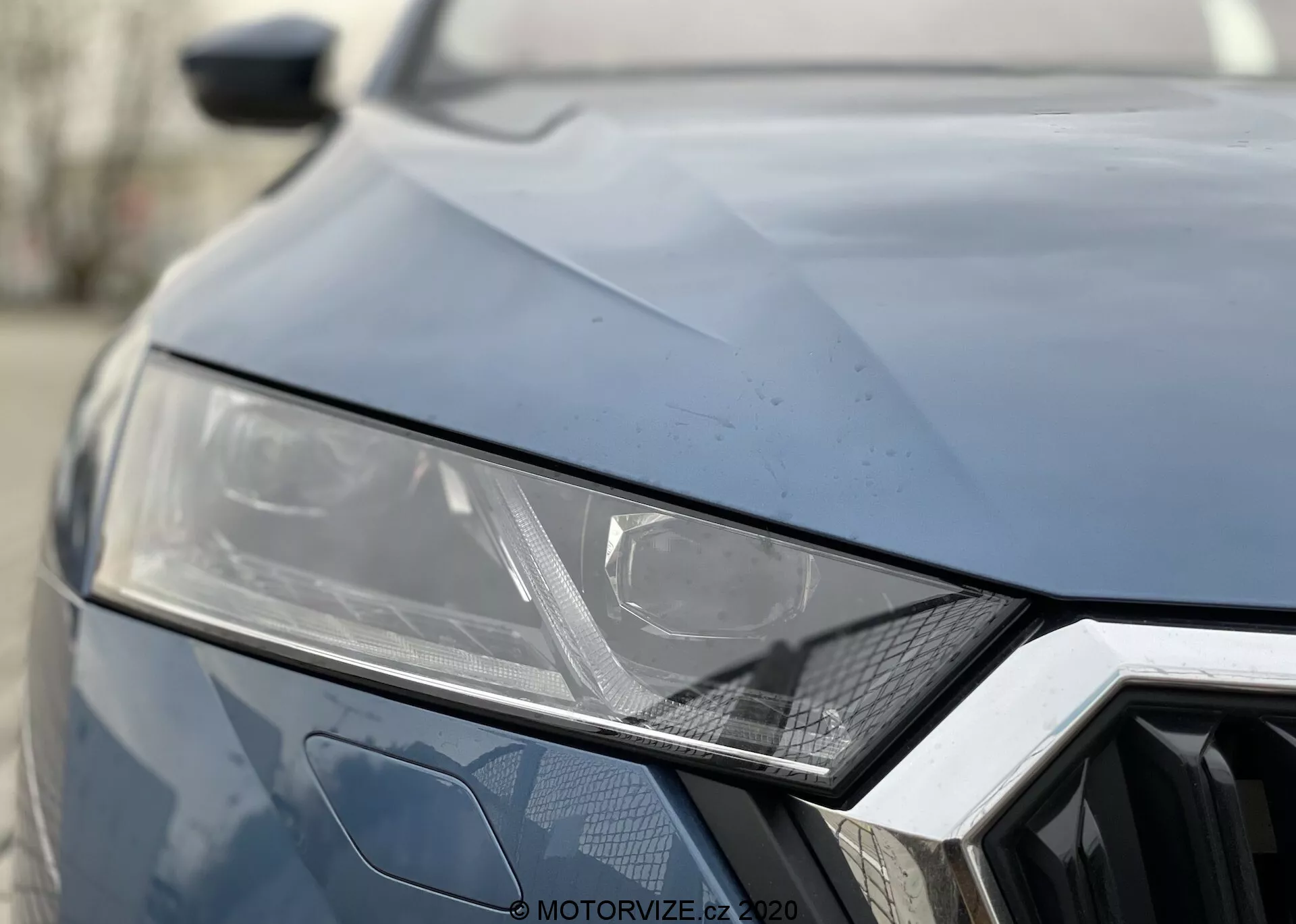 Zbliżenie przedniej części Skody Octavia 2020. Ciemny kolor, pokazujący lewą lampę przednią z eleganckim, kątowym wzornictwem sugerującym technologię LED lub HID, geometryczny sześciokątny wzór kratki i kątowe przetłoczenia na masce dla sportowego wyglądu.