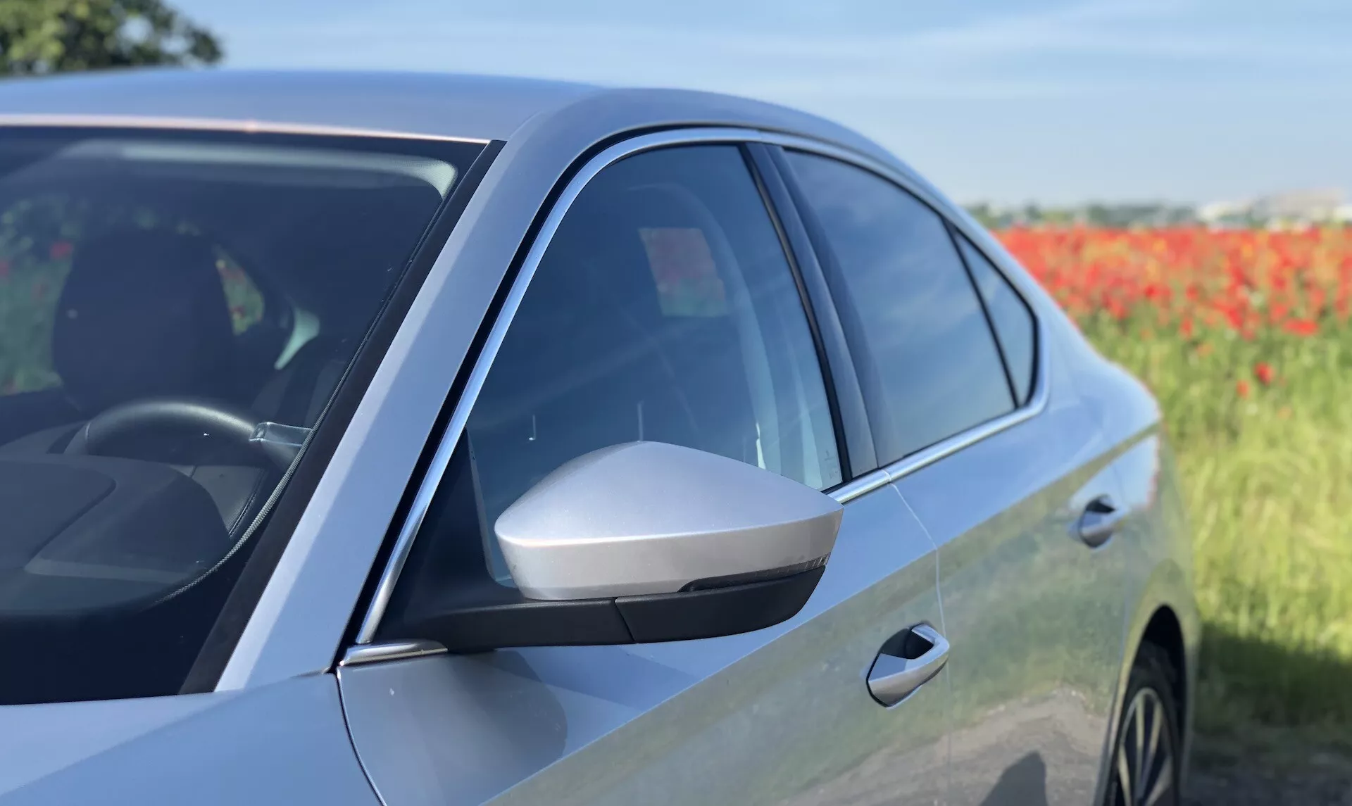 Zbliżony widok boczny nowoczesnego sedana Škoda Octavia 2020, ukazujący przednie drzwi kierowcy i lusterko boczne. Zdjęcie podkreśla opływowe lusterko boczne z wbudowanym kierunkowskazem, przyciemniane szyby, błyszczące wykończenie lakieru i aerodynamiczny design od środka do przodu. Odbicie na panelu drzwi i części przedniego nadkola sugerują obecność stopów aluminiowych. Fotografia podkreśla zaawansowane cechy konstrukcyjne i potencjalne technologie pojazdu na lekko rozmytym naturalnym tle.