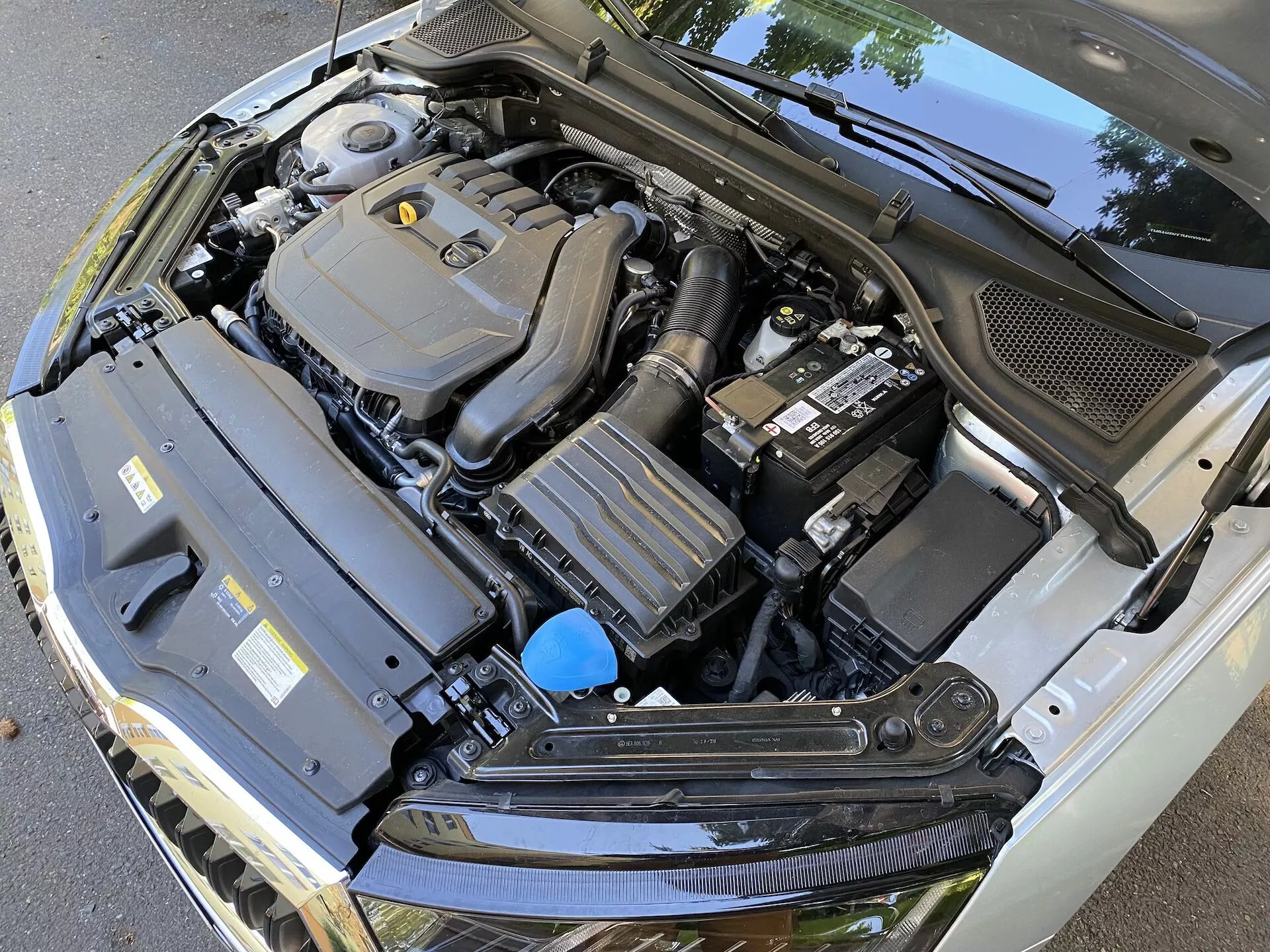 Zbliżenie komory silnika Skoda Octavia 2020 limousine z otwartą maską, pokazujące różne komponenty, w tym centralnie umieszczoną pokrywę silnika, akumulator po prawej stronie, prostokątny komponent z żeberkami chłodzącymi po lewej stronie i zbiornik płynu z niebieskim korkiem. Zdjęcie, wykonane z opadającego, lekko przesuniętego kąta, również pokazuje czarną plastikową osłonę na dole przedniej szyby, podkreślając techniczną złożoność komory silnika w samochodzie.