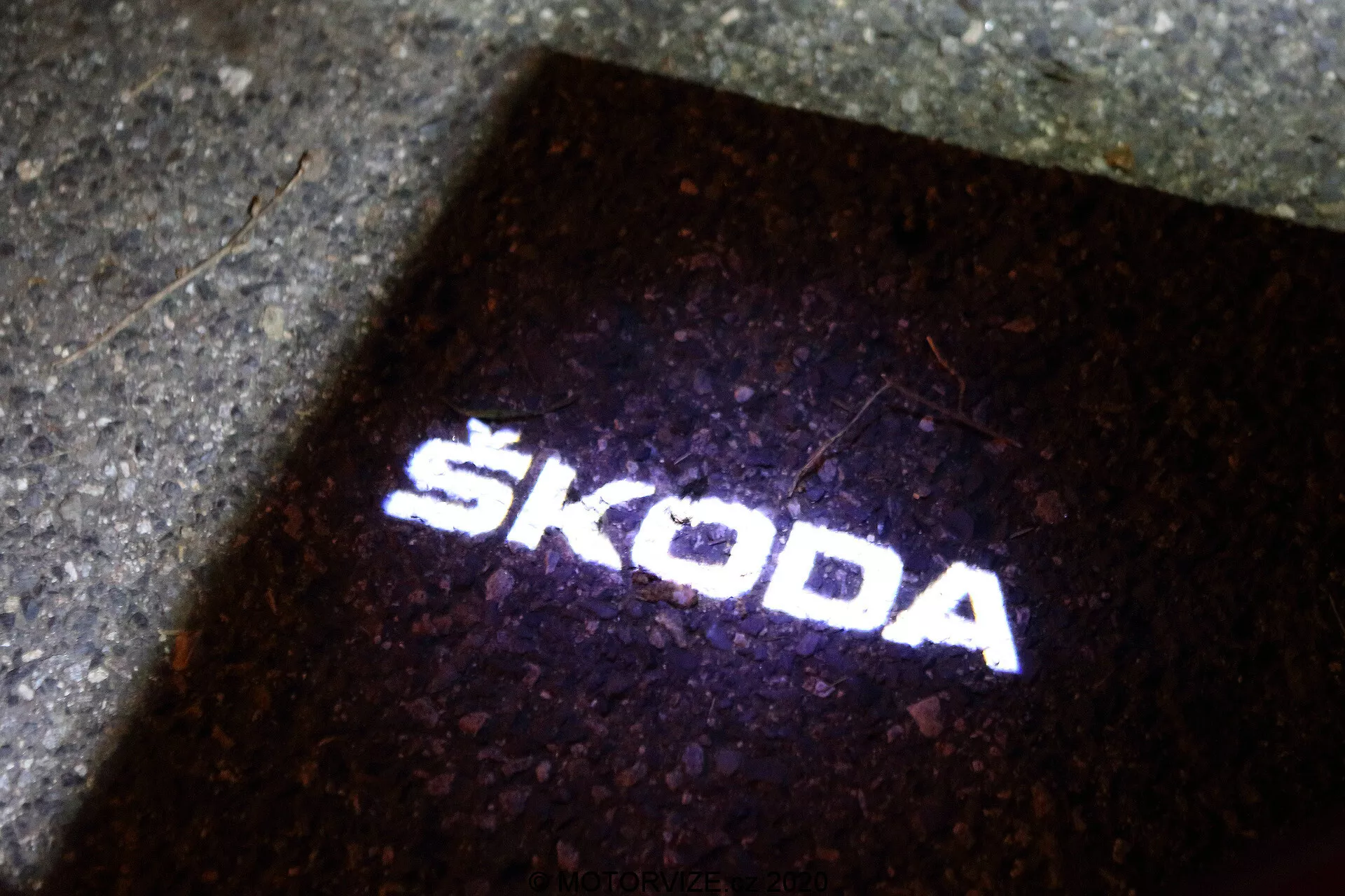 Projekcja logo Škoda na ziemi. Źródło światła, prawdopodobnie umieszczone na zewnątrz samochodu Škoda Octavia 2020 (octavia-limousine), tworzy 'puddle light', który oświetla ziemię po otwarciu drzwi pojazdu. Ta ostra i wyraźna biała projekcja światła na ciemnym tle służy zarówno celom funkcjonalnym, jak i brandingowym, co wskazuje na obecność nowoczesnej technologii LED w akcesoriach pojazdu.