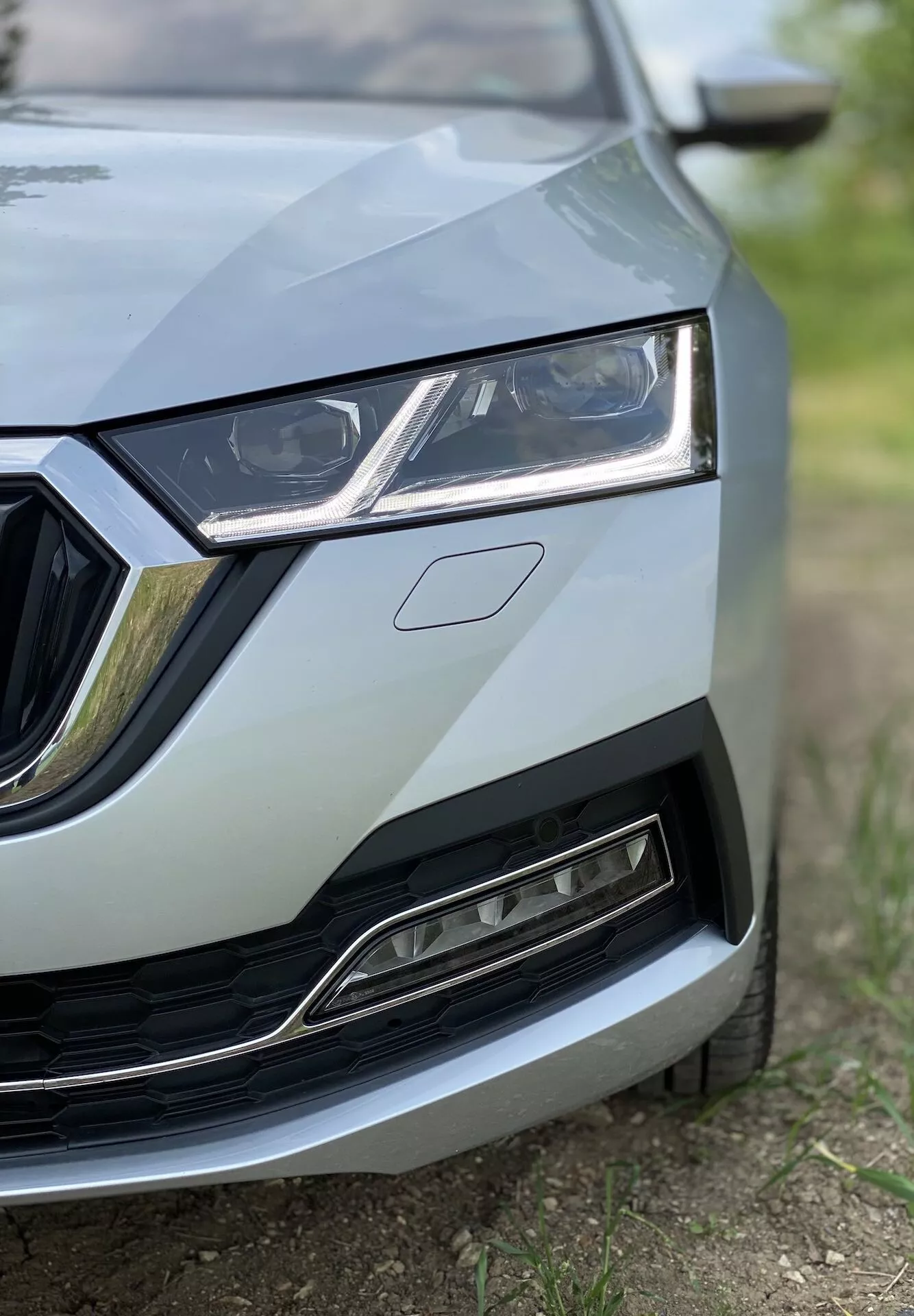 Zbliżenie przedniej lewej części Skody Octavia 2020 (Octavia Limousine) prezentujące szczegóły od środka kraty do lewego reflektora, część maski i fragment przedniego zderzaka oraz błotnika. Obraz wykonany z niskiego kąta podkreśla elegancki design reflektora z LED-owymi światłami do jazdy dziennej, kratę z chromowanymi akcentami i dolną kratę z nowoczesnym, metalicznym wykończeniem. Skupienie jest na szczegółach samochodu przy niskiej głębi ostrości, tworząc profesjonalny, wysokiej jakości wygląd.
