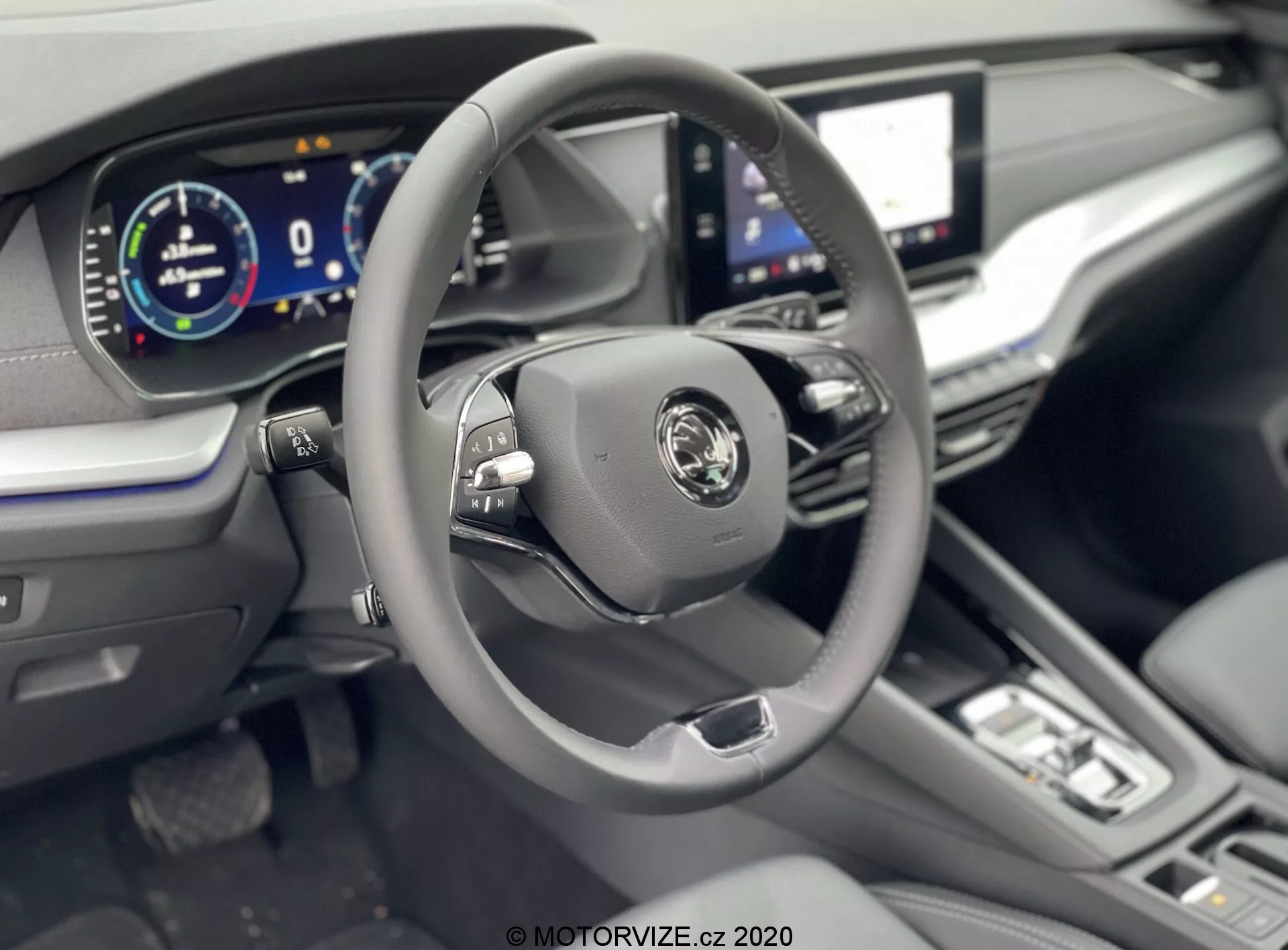 Wnętrze Skody Octavii Limousine 2020 koncentrujące się na obszarze kierowcy z lekko podwyższonego kąta, ukazujące nowoczesną trójramienną kierownicę z przyciskami wielofunkcyjnymi, centralnym logo producenta i w pełni cyfrowym zestawem wskaźników pokazującym 0 km/h. Widoczne elementy obejmują duży wyświetlacz dotykowy infotainment, minimalistyczne przyciski i pokrętła do sterowania klimatyzacją, dźwignię zmiany biegów wskazującą na automatyczną skrzynię biegów, oraz skórzaną kierownicę, skórzaną tapicerkę i chromowane akcenty dla poczucia luksusu