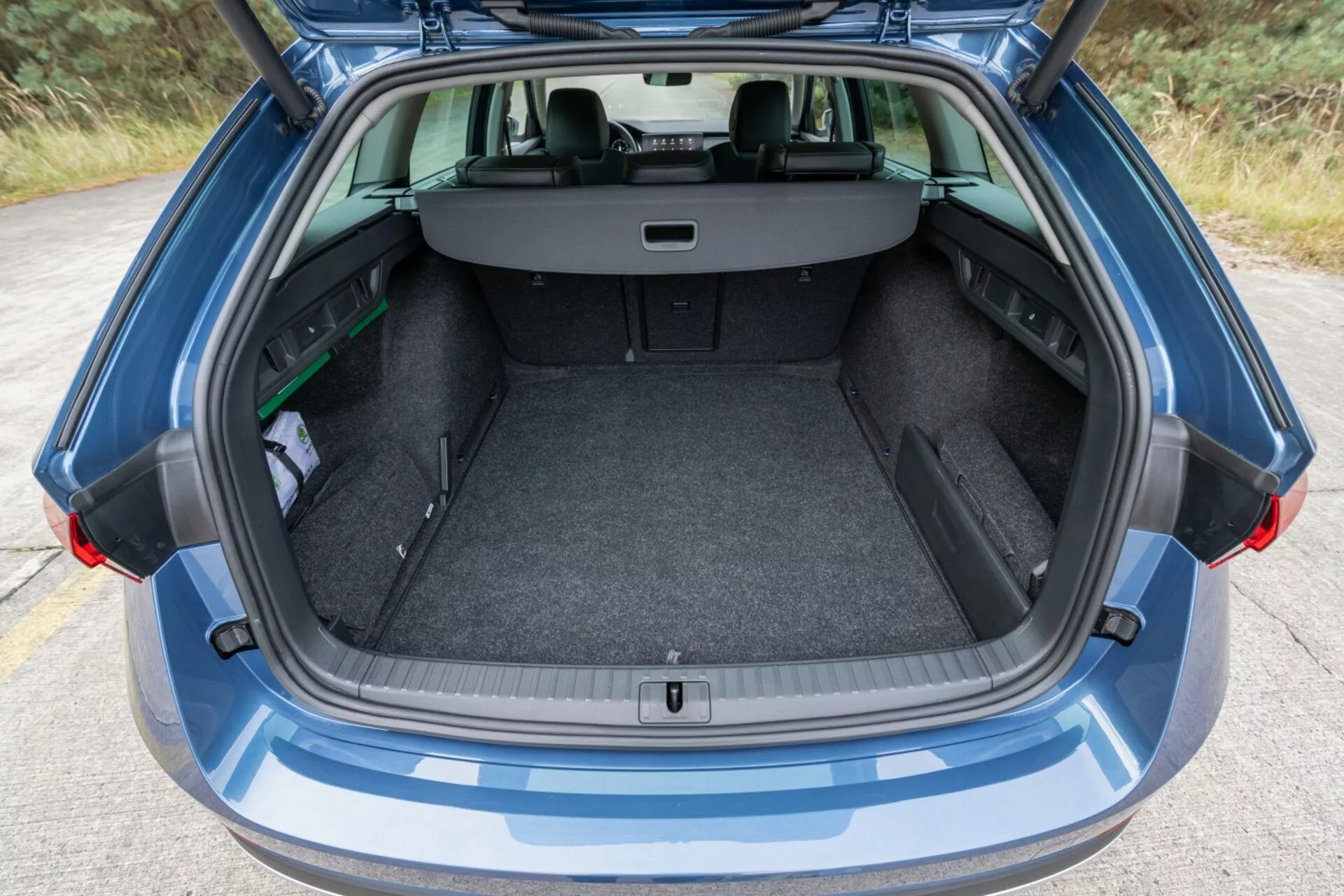 Tylny widok Skoda Octavia Limousine 2017 z podkreśleniem jej przestronnego, czystego bagażnika z otwartą klapą bagażnika, rozsuwaną pokrywą bagażu i widoczną konfiguracją tylnych siedzeń w proporcji 60/40 dla zwiększonej pojemności bagażowej, obok małego parasola i zielonego przedmiotu, w świetle otoczenia.