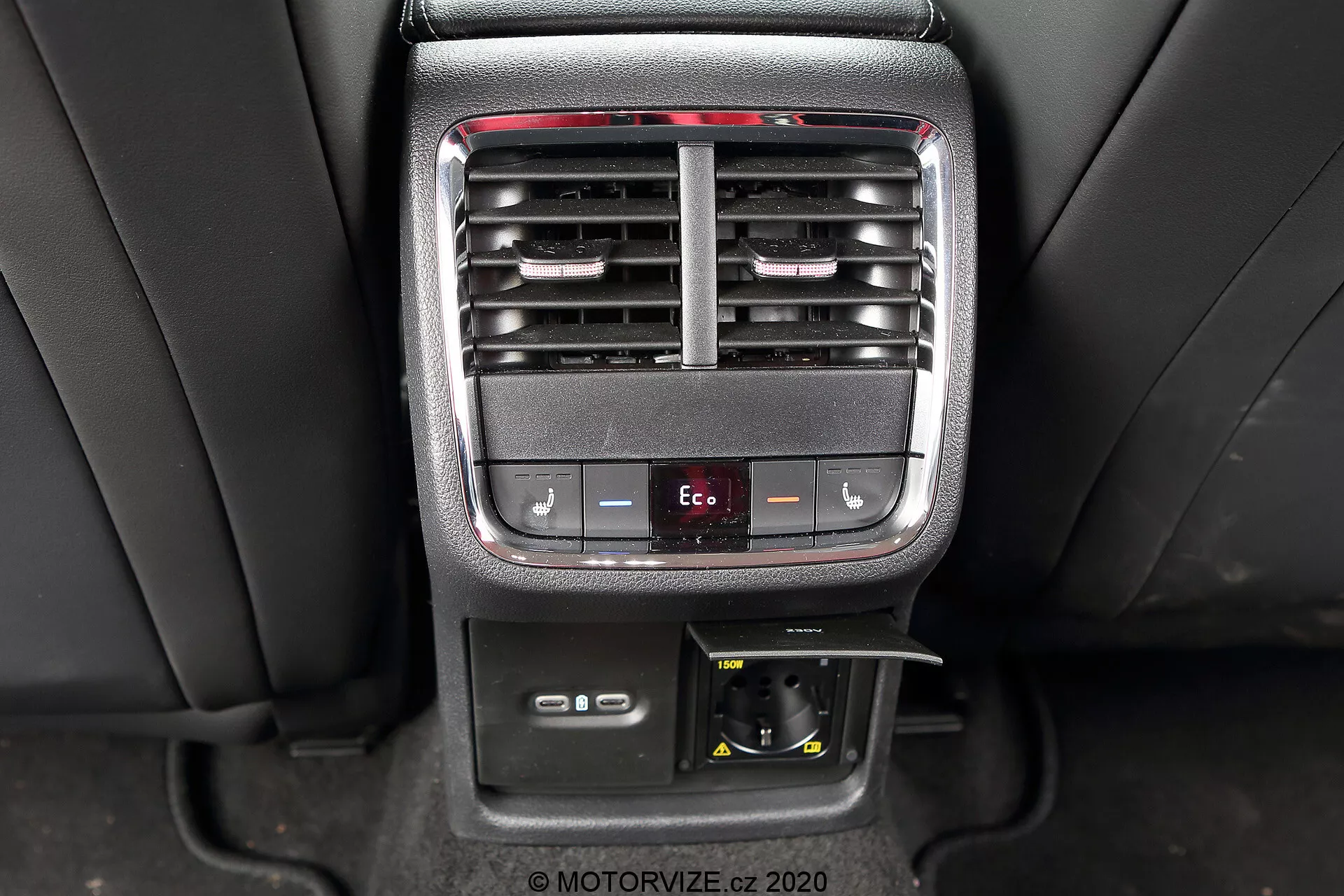 Obraz przedstawia środkową konsolę wnętrza Skody Octavia Limousine 2020, podkreślając cechy kontroli klimatu i wygody dla pasażerów tylnych. Zdjęcie wyraźnie ukazuje regulowane kratki wentylacyjne, przyciski do regulacji temperatury, przycisk trybu Eco, gniazdo zasilania 12V i schowek.
