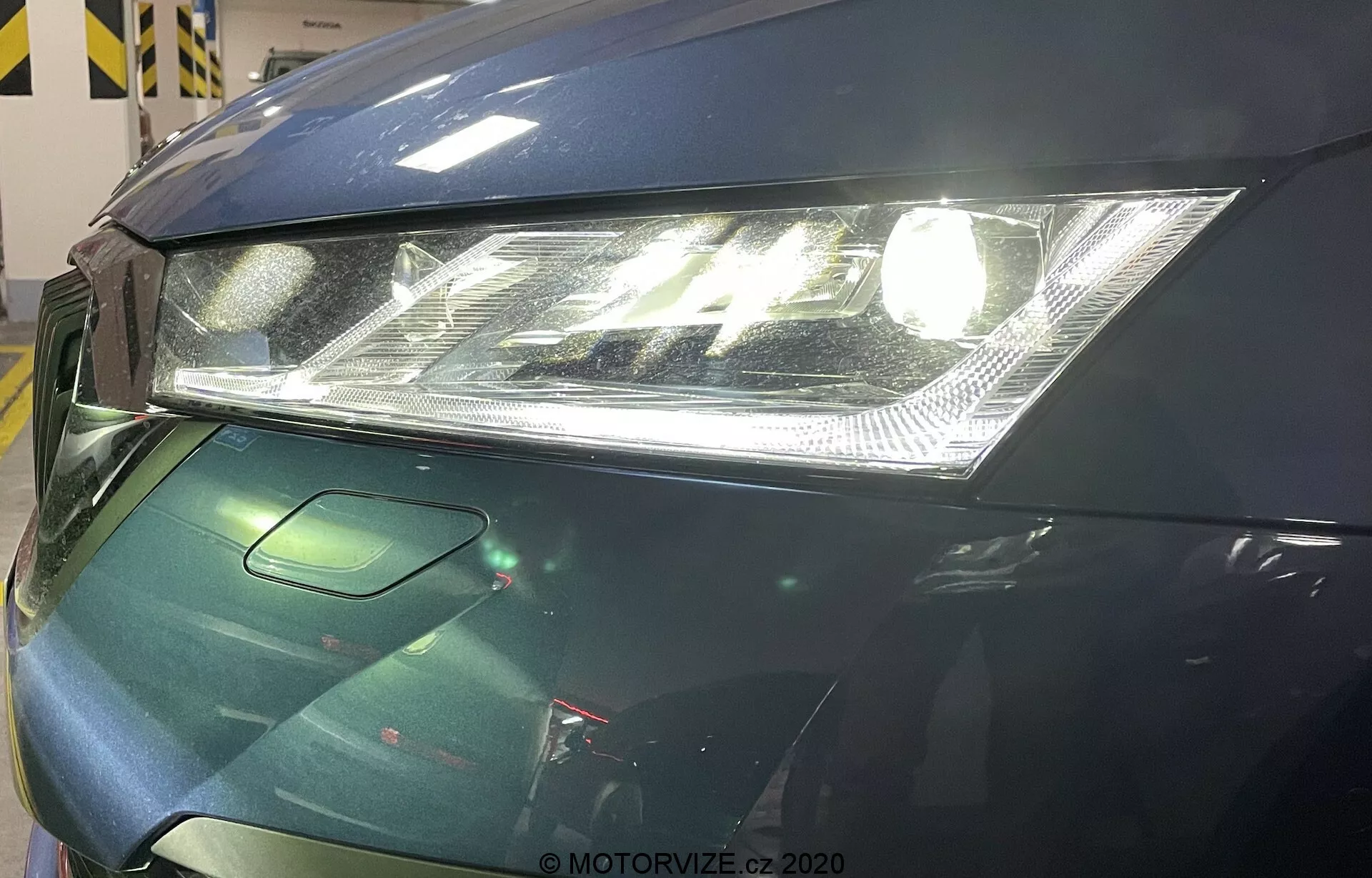 Zbliżenie przedniej lewej części Skody Octavia 2020. Obraz koncentruje się na nowoczesnym reflektorze z przezroczystą soczewką i zintegrowanymi światłami do jazdy dziennej LED, częściowo widocznej masce o gładkim kształcie i małym fragmencie osłony chłodnicy, która prawdopodobnie integruje czujniki wspomagające kierowcę. Samochód ma ciemnozielony, błyszczący lakier, co sugeruje zastosowanie zaawansowanych środków ochrony lakieru.