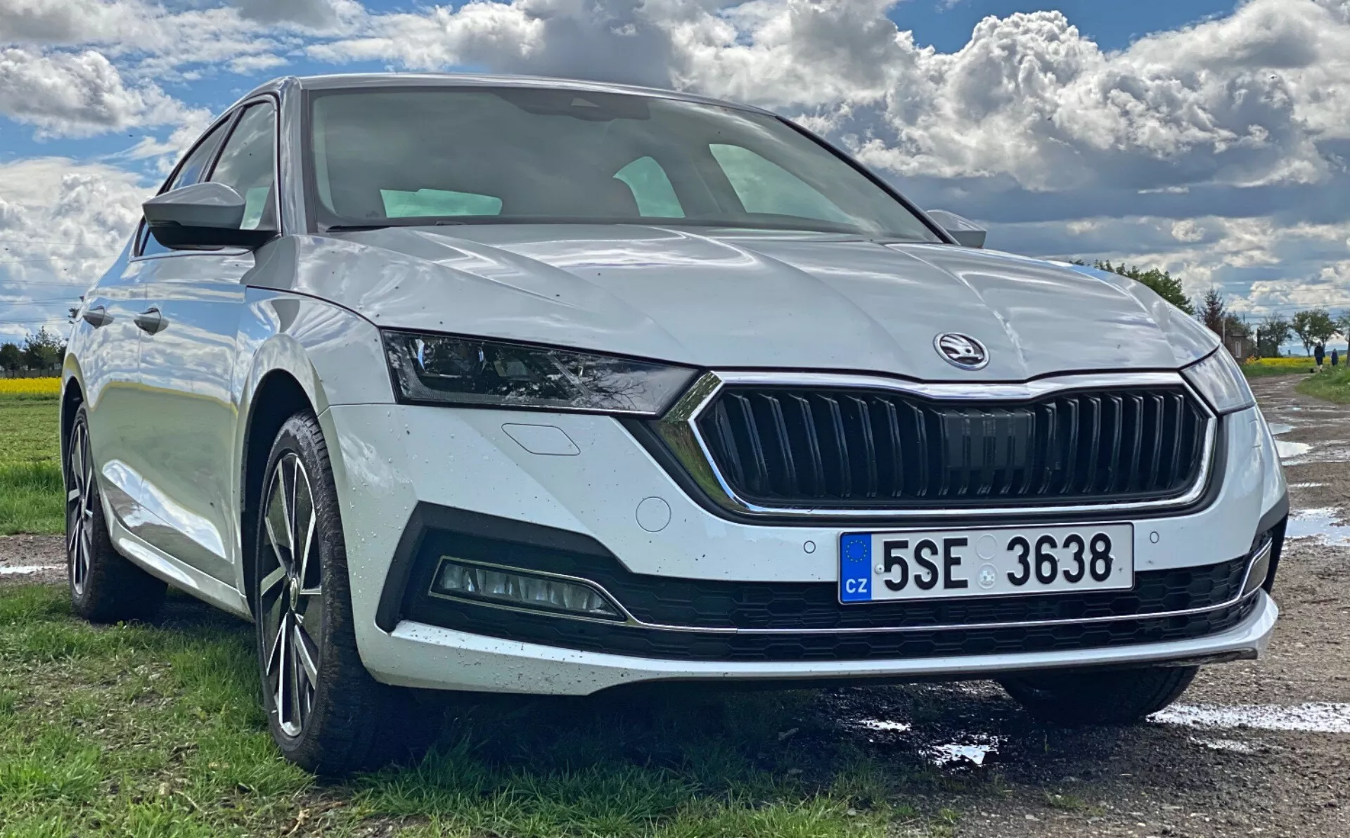 Niskokątny przedni-czwarty widok białej Škody Octavia 2020 zaparkowanej na świeżym powietrzu, podkreślający przednią fasadę, ostro zaprojektowane reflektory z diodami LED do jazdy dziennej, szeroką kratkę z logo Škoda, czujnik parkowania, sportowy przedni zderzak z reflektorami przeciwmgielnymi, aluminiowe felgi wieloramienne i zintegrowany kierunkowskaz w lusterku bocznym. Widoczne są również panoramiczna szyba czołowa i europejska tablica rejestracyjna.