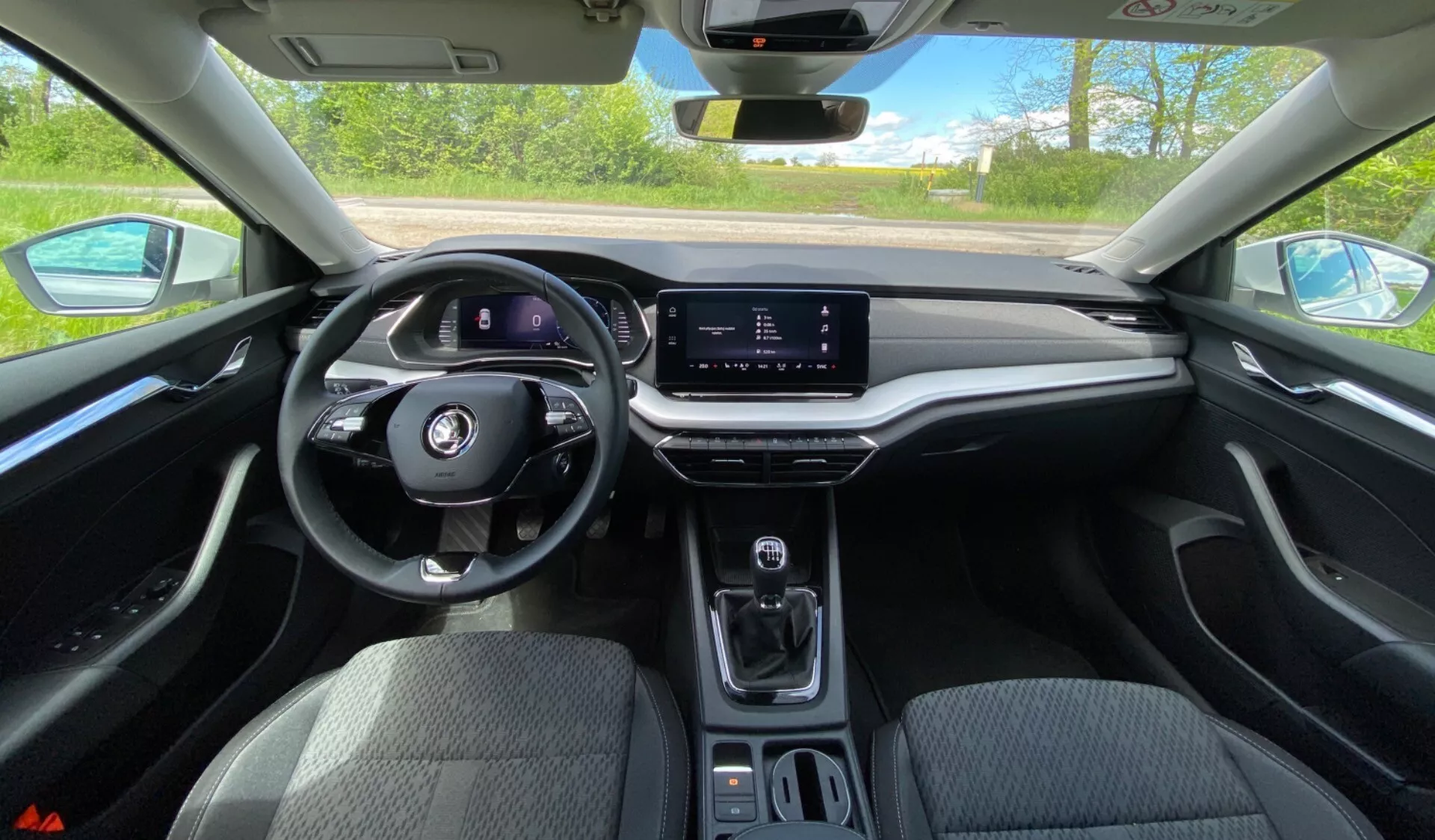 Widok wnętrza Skoda Octavia 2020 z perspektywy kierowcy, pokazujący wielofunkcyjną kierownicę, cyfrowy zestaw wskaźników, poziomy ekran dotykowy systemu multimedialnego, minimalistyczną konsolę środkową z selektorem biegów, uchwyty na kubki i miękką deskę rozdzielczą z wykończeniem przypominającym drewno w jasnym świetle dziennym.
