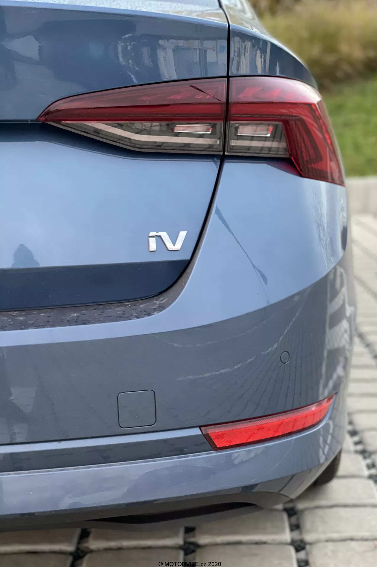 Zbliżenie tylnej części Skoda Octavia 2020 pod kątem ukośnym, skupiające się na nowoczesnym, kanciastym światle tylnym prawdopodobnie z elementami LED. Poniżej światła tylnego widoczny jest mały prostokątny odblask lub dodatkowe oświetlenie. Metaliczne nadwozie w kolorze niebiesko-szarym posiada delikatnie zakrzywioną pokrywę bagażnika, dobrze zintegrowany tylny zderzak oraz odznakę 'iV', co wskazuje na napęd hybrydowy lub elektryczny. Słabo widoczne czujniki parkowania w prawym dolnym rogu sugerują zaawansowane systemy wspomagania kierowcy. Zdjęcie, wykonane w świetle dziennym z małą głębią ostrości, pokazuje auto zaparkowane na brukowanej nawierzchni.