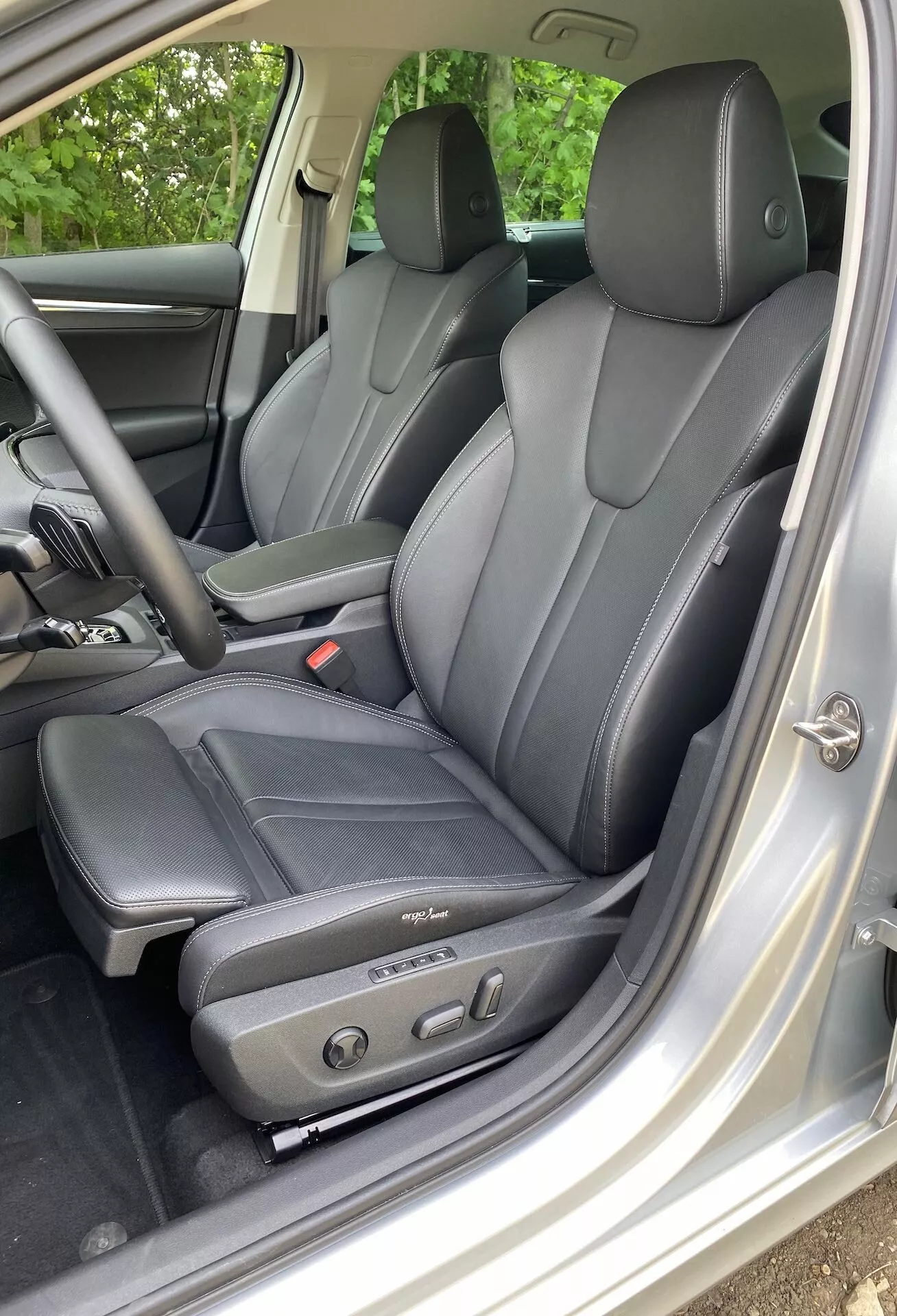 Widok wnętrza Škoda Octavia limousine 2020 skoncentrowany na obszarze kierowcy, z ciemnymi skórzanymi siedzeniami o ergonomicznym kształcie i elektrycznymi panelami sterującymi, widok od lewej strony przez otwarte drzwi kierowcy. Widoczne zagłówki dla kierowcy i pasażerów tylnych wskazują na miejsce dla co najmniej czterech osób. Wewnętrzne wykończenie w kolorze srebrnym lub jasnoszarym kontrastuje z siedzeniami, a panel drzwi zawiera elektryczne sterowanie szybami i lusterkami, podkreślając technologiczny komfort i luksus.