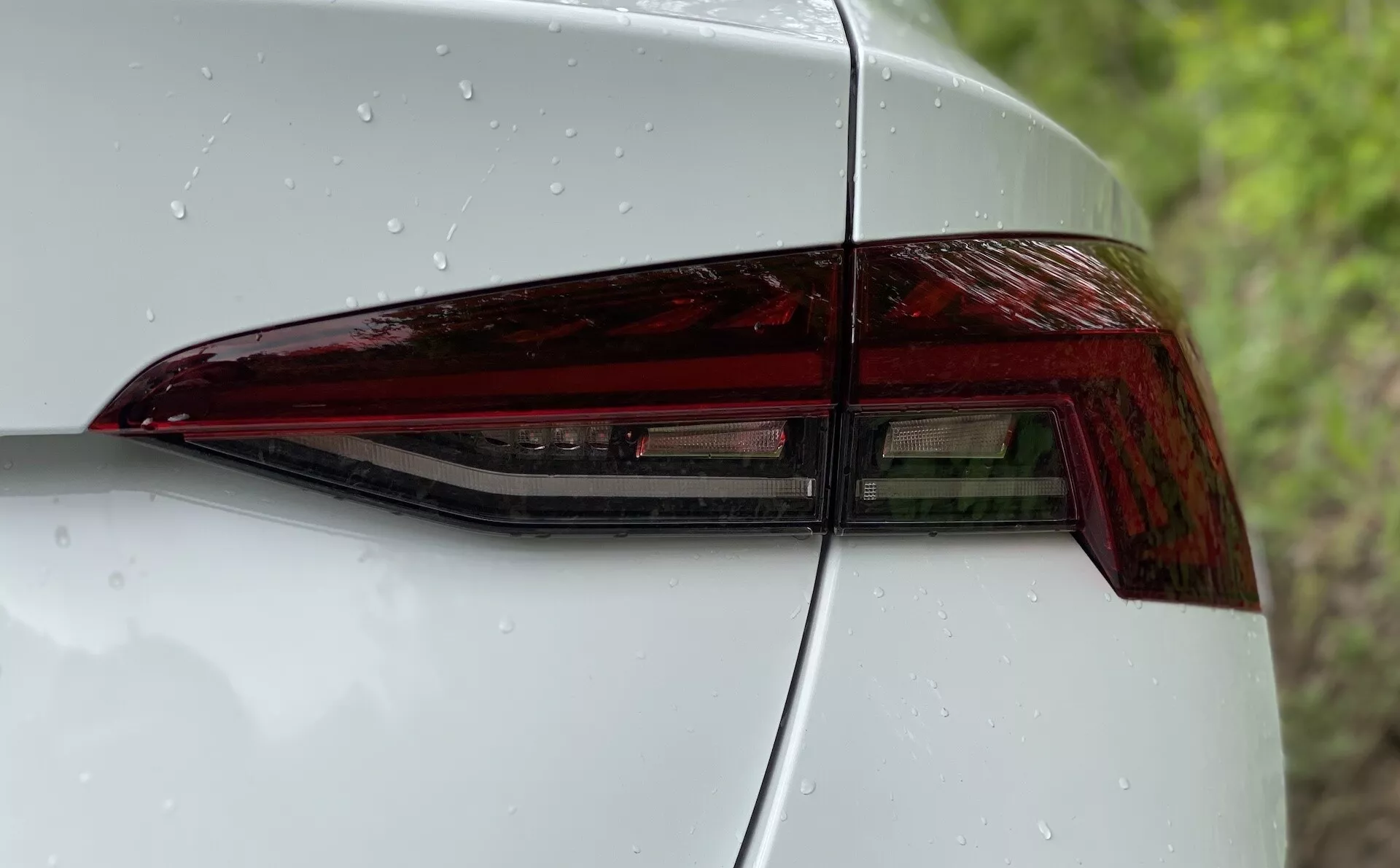 Zbliżenie na tylną lewą stronę Škoda Octavia 2020, z naciskiem na zestaw tylnych świateł. Zdjęcie wykonane jest pod kątem ukośnym, podkreślając nowoczesny design tylnego światła LED z ostrymi kątami i błyszczącym wykończeniem. Widać część tylnego panelu i pokrywę bagażnika, co pokazuje bezszwową integrację i estetykę aerodynamiczną. Gładka powierzchnia z kroplami wody wskazuje na niedawne opady lub świeże mycie.