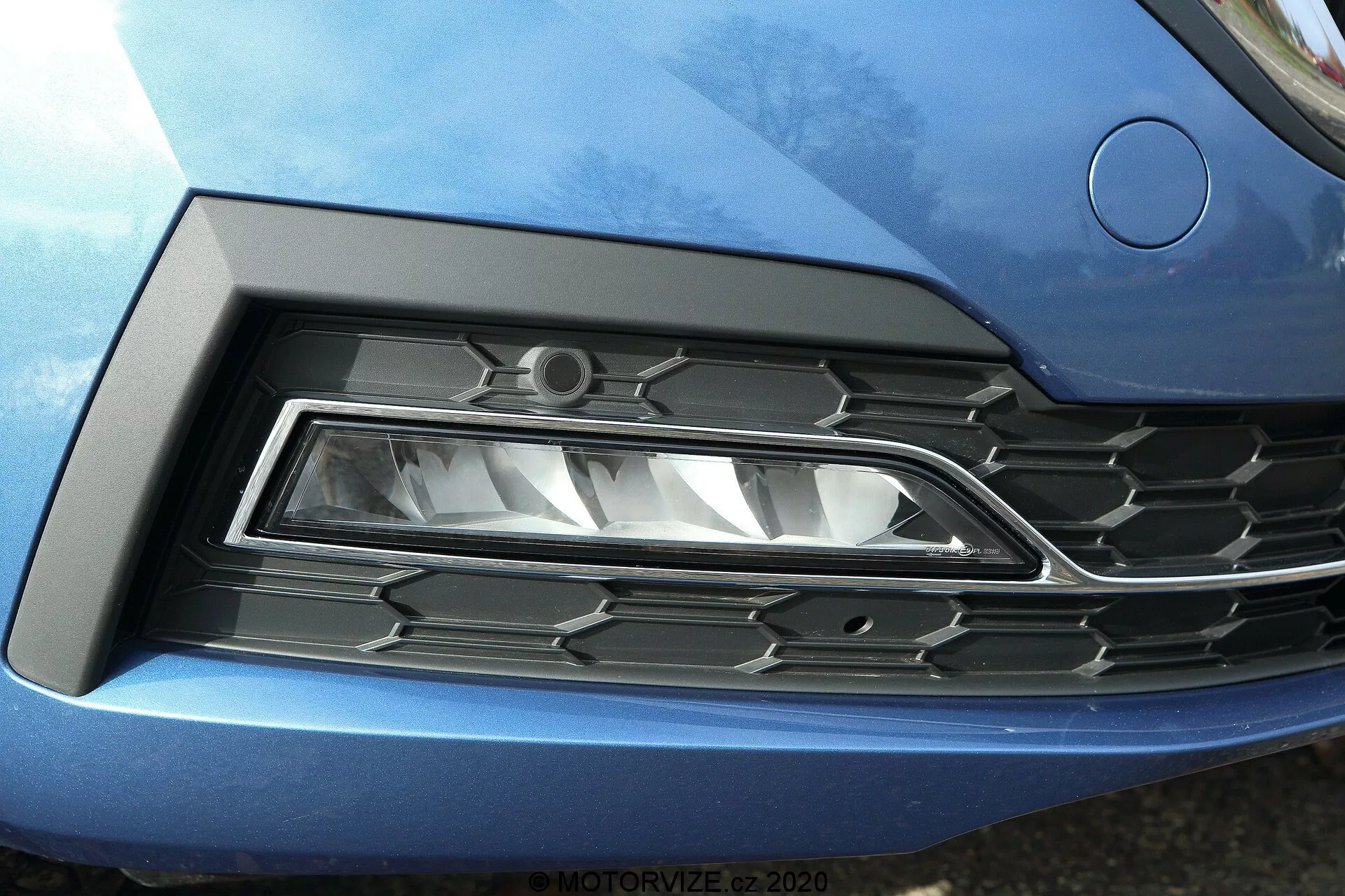 Zbliżenie przedniego detalu Skody Octavii 2020 (Octavia Limousine), z naciskiem na lewy reflektor i kratkę. Nowoczesny reflektor LED o ostrym, kanciastym designie jest umieszczony w rzeźbionym wgłębieniu, które płynnie przechodzi w metaliczne, niebieskie nadwozie samochodu. Sąsiednia kratka ma wzór heksagonalny z czarnego plastiku z chromowanymi akcentami, odzwierciedlając współczesny styl motoryzacyjny.