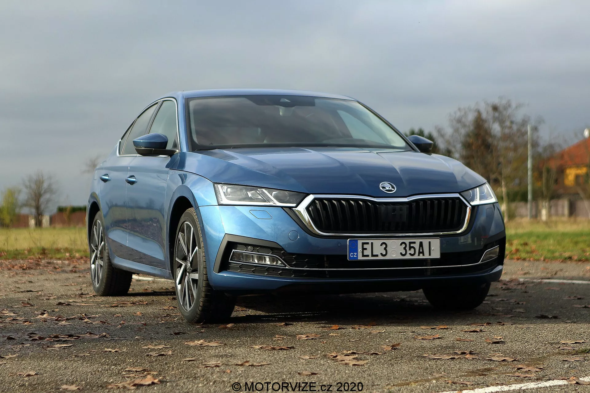 Obraz niebieskiej Škody Octavia Limousine 2020 uchwycony z niskiego, przedniego trzech czwartych kąta, podkreślający charakterystyczną grill przedni z chromowanym konturem, ostre reflektory LED, konturowaną maskę, nowoczesne felgi aluminiowe oraz eleganckie lusterka boczne z zintegrowanymi kierunkowskazami. Gładka karoseria i lekko przyciemniane bezramkowe okna sugerują wyższy poziom wykończenia, a cała długość samochodu jest widoczna, w tym odznaki wskazujące model lub typ silnika.