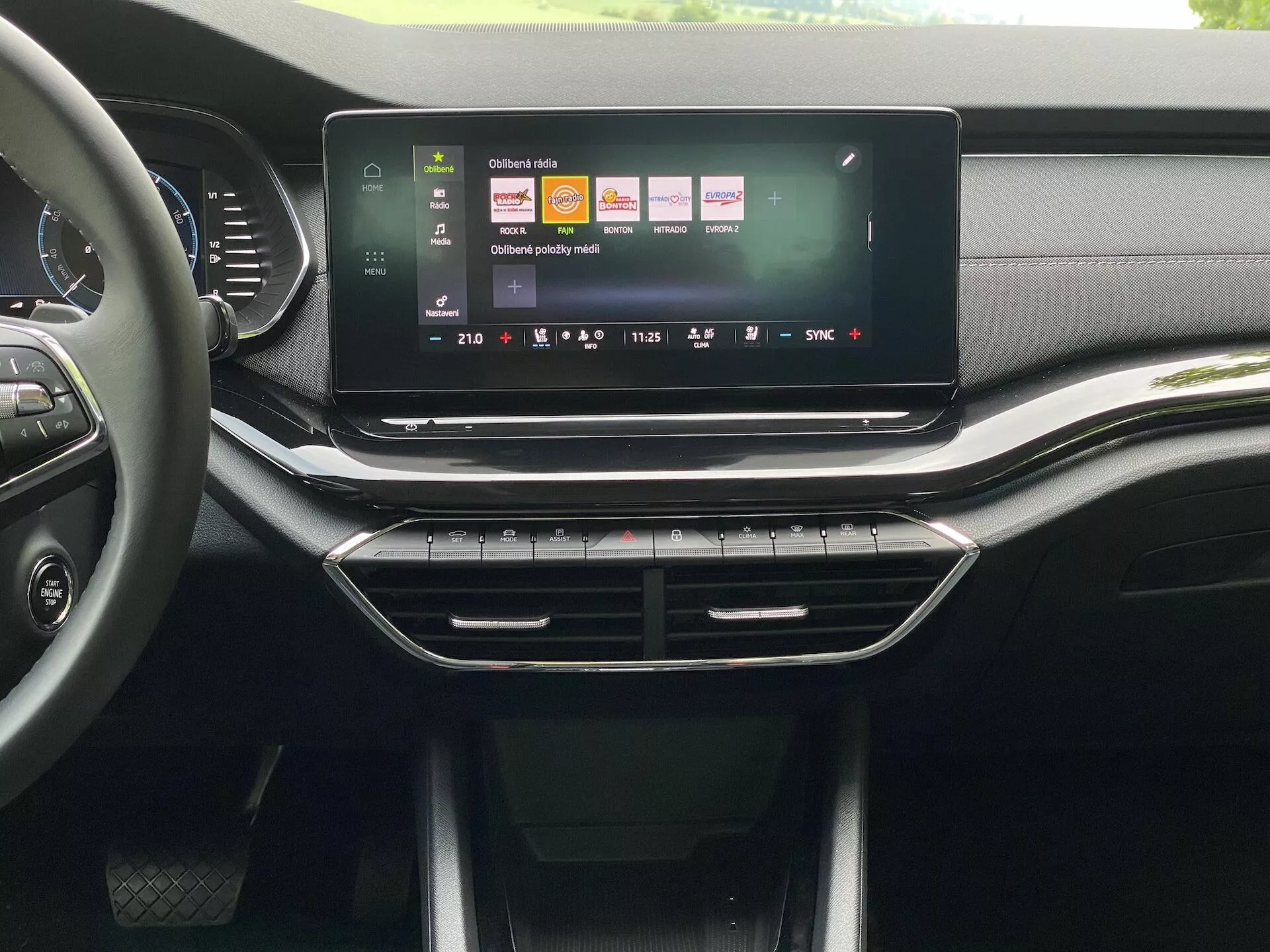 Wnętrze Škoda Octavia 2020 sedan, z wyróżnieniem centralnej konsoli i systemu infotainment z perspektywy przedniego siedzenia. Duży, panoramiczny ekran dotykowy ukazuje barwny interfejs użytkownika z ikonami sugerującymi funkcjonalności takie jak nawigacja, odtwarzanie mediów i ustawienia pojazdu. Pod ekranem znajdują się uproszczone kratki nawiewu z chromowanym wykończeniem, a następnie fizyczne przyciski i pokrętła do sterowania klimatyzacją. Tablica rozdzielcza i konsola mają mieszankę tekstur przypominających skórę oraz matowych i błyszczących plastików, co sugeruje wysoką jakość wykończenia w średnim do wyższego segmencie rynku.