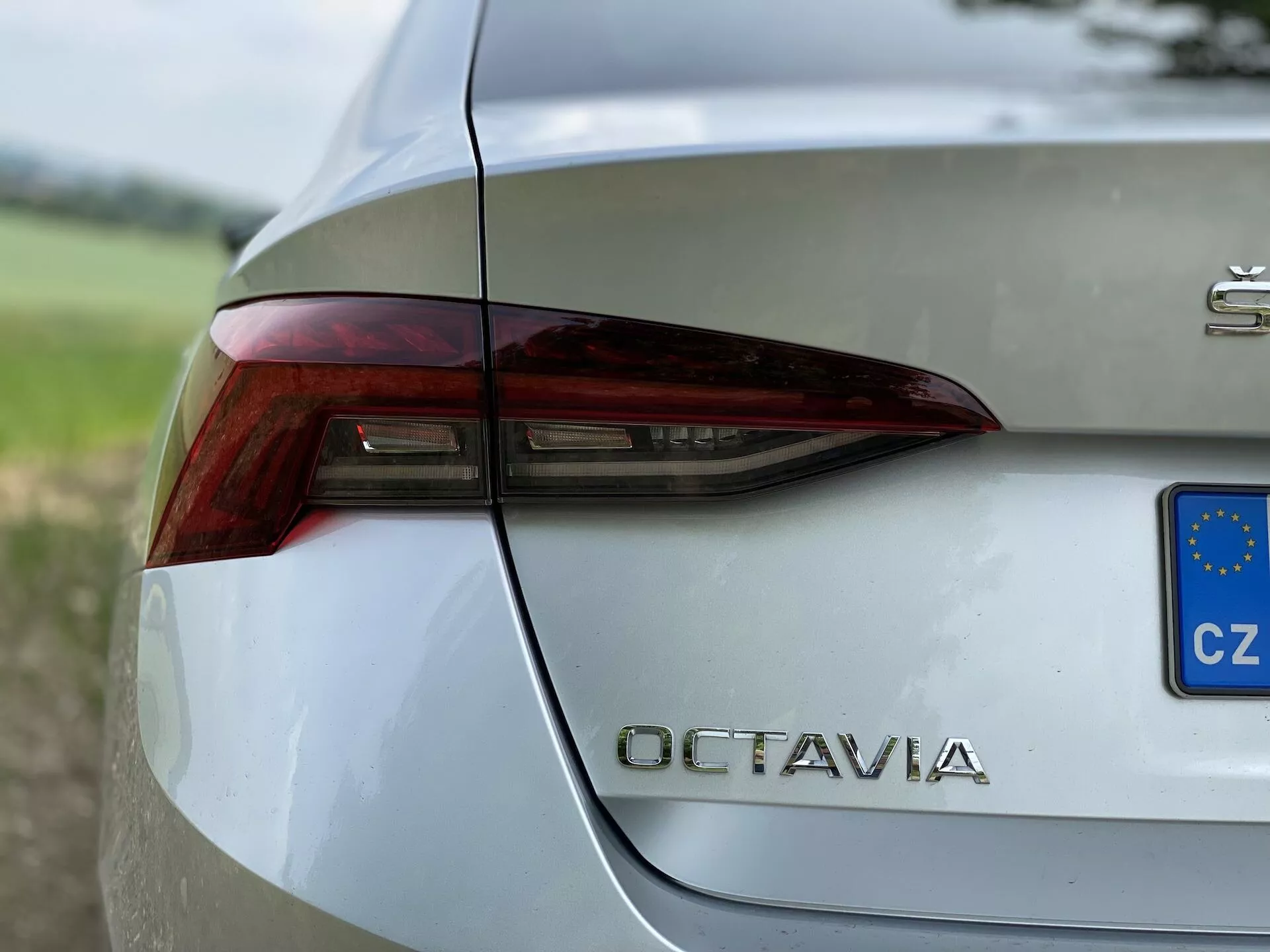 Zbliżenie tylnej prawej części srebrnego Škoda Octavia 2020. Obraz uchwyca ostre i kanciaste światła tylne z zintegrowanymi elementami dla świateł hamowania, kierunkowskazów i prawdopodobnie świateł cofania. Klapa bagażnika wyraźnie pokazuje oznaczenie modelu 'OCTAVIA' chromowanymi literami poniżej tylnej lampy. Mały niebiesko-żółty prostokąt z gwiazdkami wskazuje na rejestrację w Republice Czeskiej. Polerowane, metaliczne wykończenie nadwozia odbija otoczenie, ustawione na rozmytym tle zieleni, podkreślając jego wyrafinowany design.