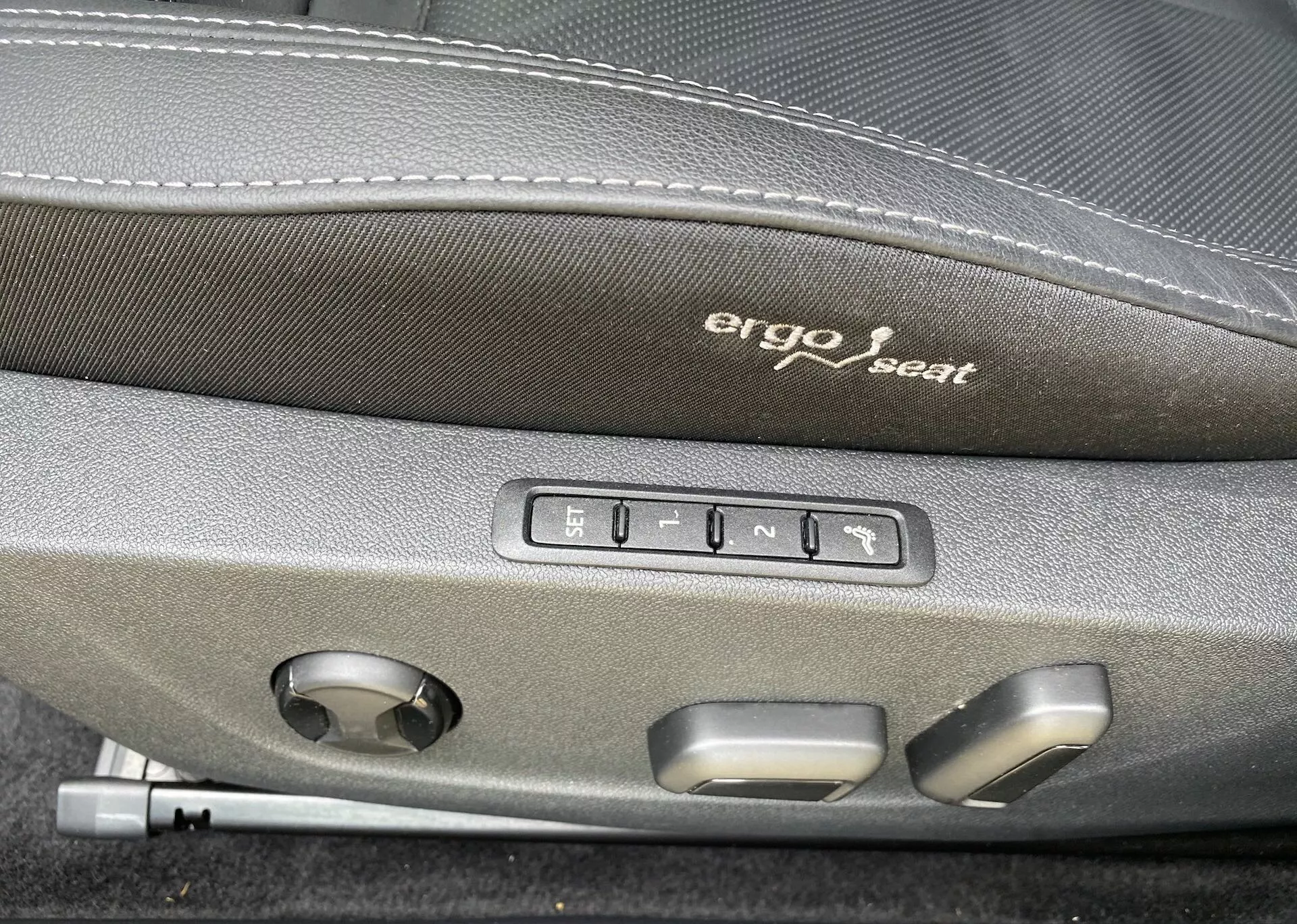 Zbliżenie wnętrza Škody Octavii 2020, skupiające się na ergonomicznych przyciskach do regulacji siedzenia. 'Ergo seat' jest wyhaftowane na ciemniejszej górnej części siedzenia. Poniżej haftu widać pięć przycisków: jeden do ustawień pamięci siedzenia i cztery do regulacji siedzenia, takich jak do przodu, do tyłu, w górę, w dół i kąt nachylenia oparcia. Jest też okrągłe pokrętło, prawdopodobnie służące do regulacji podparcia lędźwiowego lub innych wielofunkcyjnych regulacji siedzenia, co wskazuje na wyższy komfort i zaawansowaną technologię w tym pojeździe.