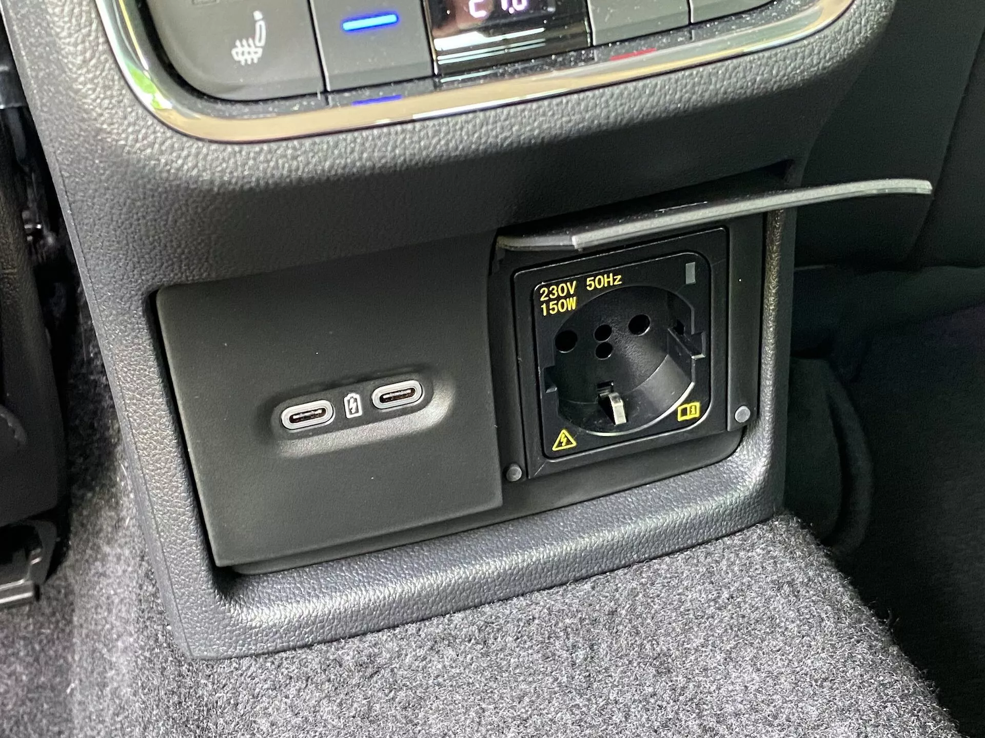 Zbliżenie wnętrza Skoda Octavia 2020 z funkcjami zasilania: dolna część konsoli środkowej z dwoma portami USB-A po lewej stronie do ładowania lub podłączania do systemu multimedialnego oraz gniazdem 230V AC dla wtyczek europejskich o maksymalnej mocy 150W po prawej stronie, otoczona teksturowanym wykończeniem i widoczną wykładziną samochodową.