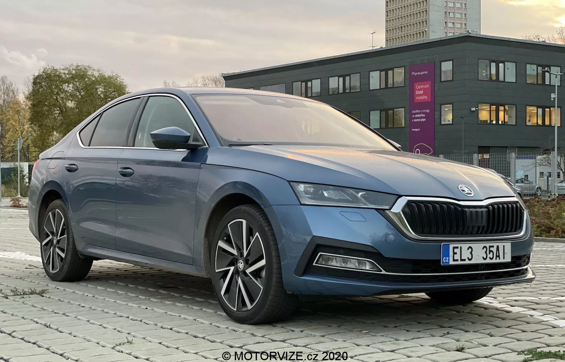 Profesjonalne zdjęcie czterodrzwiowego Skoda Octavia Limousine 2020 wykonane z przedniego lewego trzy-czwartego kąta, wyraźnie pokazujące charakterystyczną kratkę, eleganckie reflektory LED, przedni zderzak z możliwymi światłami przeciwmgielnymi, gładkie linie nadwozia, wieloszprychowe felgi aluminiowe, lusterka boczne z wbudowanymi kierunkowskazami oraz chromowaną obwódkę szyb w ekskluzywnym wykończeniu w mieście lub na przedmieściach o świcie lub zmierzchu.