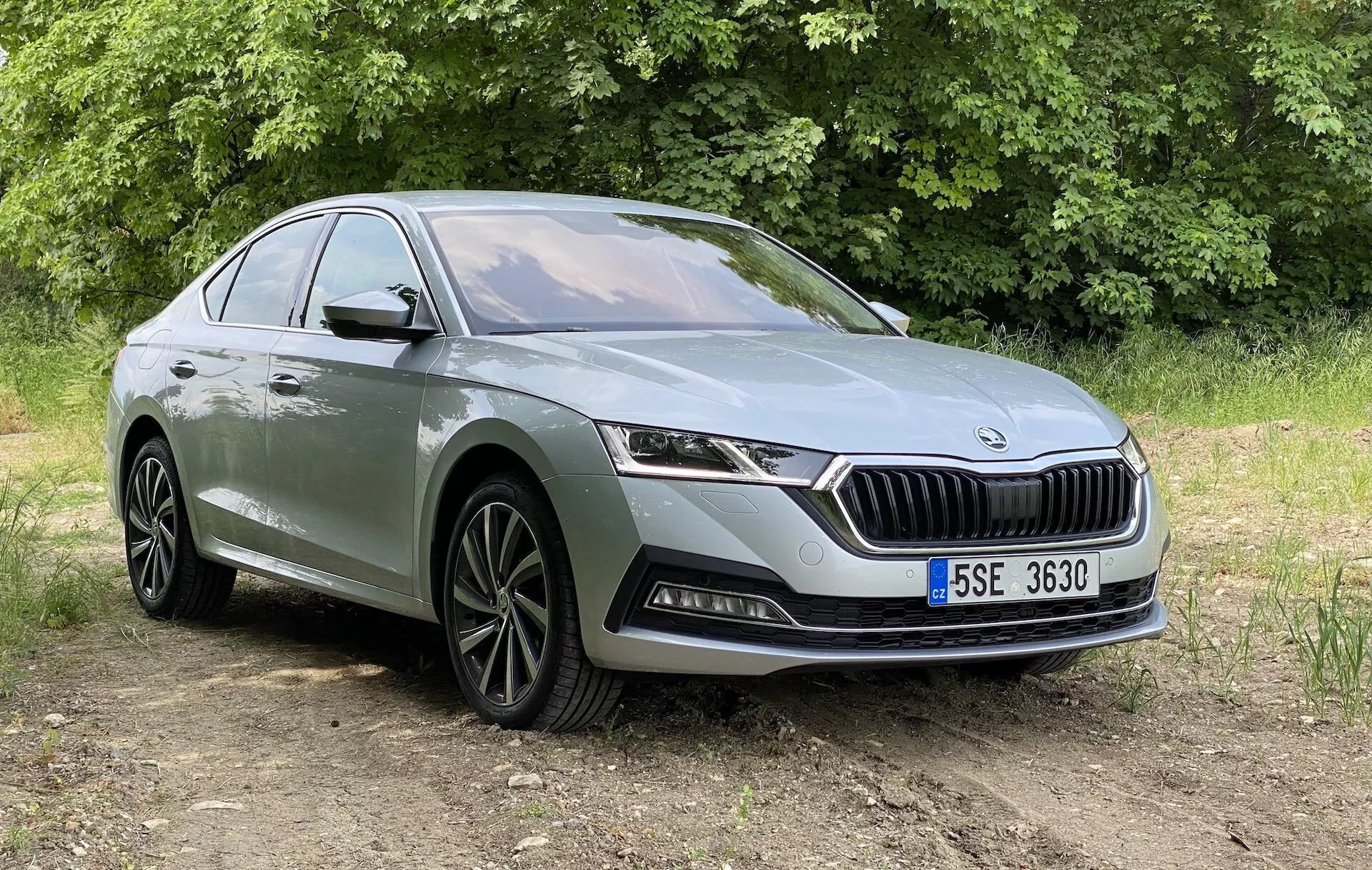 Widok przedni w trzech czwartych nowoczesnego sedana Skoda Octavia 2020, pokazany z nieco niższego kąta, koncentrujący się na przedniej części, grillu z pionowymi listwami i wyrazistym logo, eleganckich poziomych reflektorach LED oraz dolnym zderzaku z światłami przeciwmgielnymi i wlotami powietrza. Samochód jest ustawiony na nieutwardzonej powierzchni z roślinnością w tle, w naturalnym świetle dziennym. Inne widoczne szczegóły to felgi aluminiowe z wieloszprychowym wzorem, charakterystyczna linia biegnąca wzdłuż profilu bocznego i klamki drzwi. Odpowiedni do prezentacji wzornictwa samochodowego i nowoczesnych funkcji pojazdów w otoczeniu zewnętrznym.