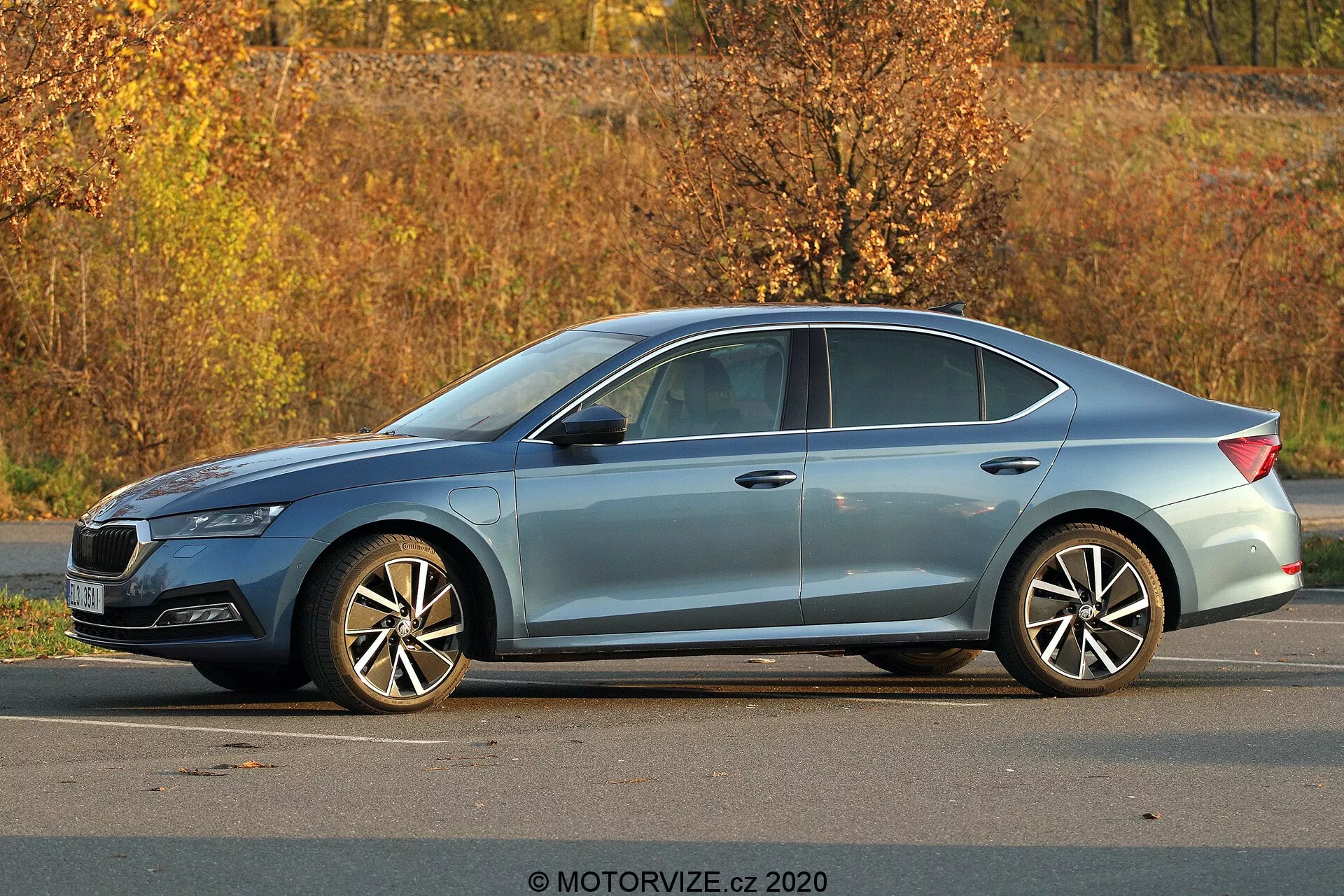 Nowoczesny sedan Skoda Octavia 2020 zaparkowany na zewnątrz, widok z boku pod kątem trzy czwarte, pokazujący pełny profil, długość i wysokość; widoczne detale obejmujące ostre reflektory, opadającą linię dachu, linie charakteru, chromowane obramowania okien, wieloramienne alufelgi, jesienny krajobraz i naturalne oświetlenie dzienne.
