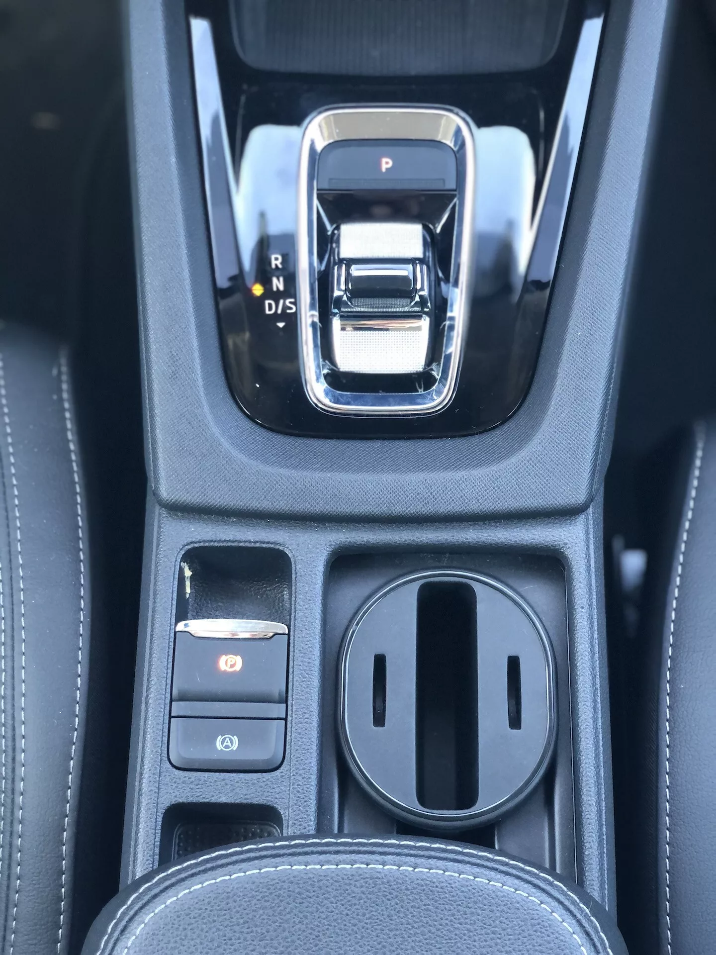Zbliżenie na obszar automatycznej zmiany biegów w Škodzie Octavia Limousine 2020. Dźwignia zmiany biegów to srebrno-czarna elektroniczna dźwignia z opcjami P, R, N i D/S. Po lewej stronie znajdują się dwa przyciski sterujące, z których górny jest podświetlony na czerwono, prawdopodobnie elektroniczny hamulec postojowy. Po prawej stronie znajduje się zamknięty schowek z dwoma uchwytami na kubki. Czarna konsola ma teksturę przypominającą skórę z detalicznym przeszyciem, co wskazuje na luksusowe wnętrze z zaawansowanymi systemami transmisji i elektronicznymi systemami parkowania.